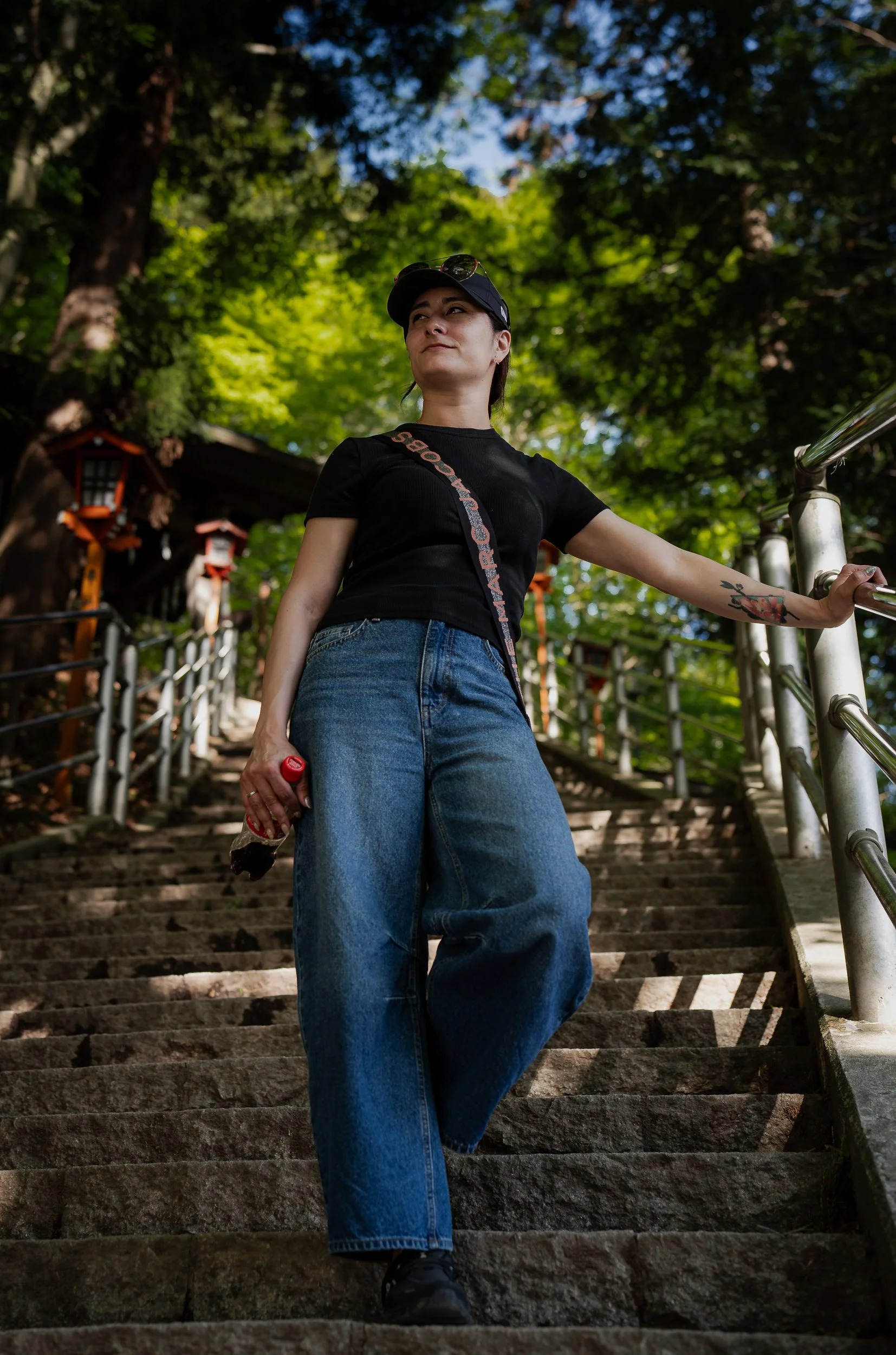 A woman wearing a black cap, black t-shirt, and blue jeans walking down stone steps outdoors surrounded by trees and green foliage.