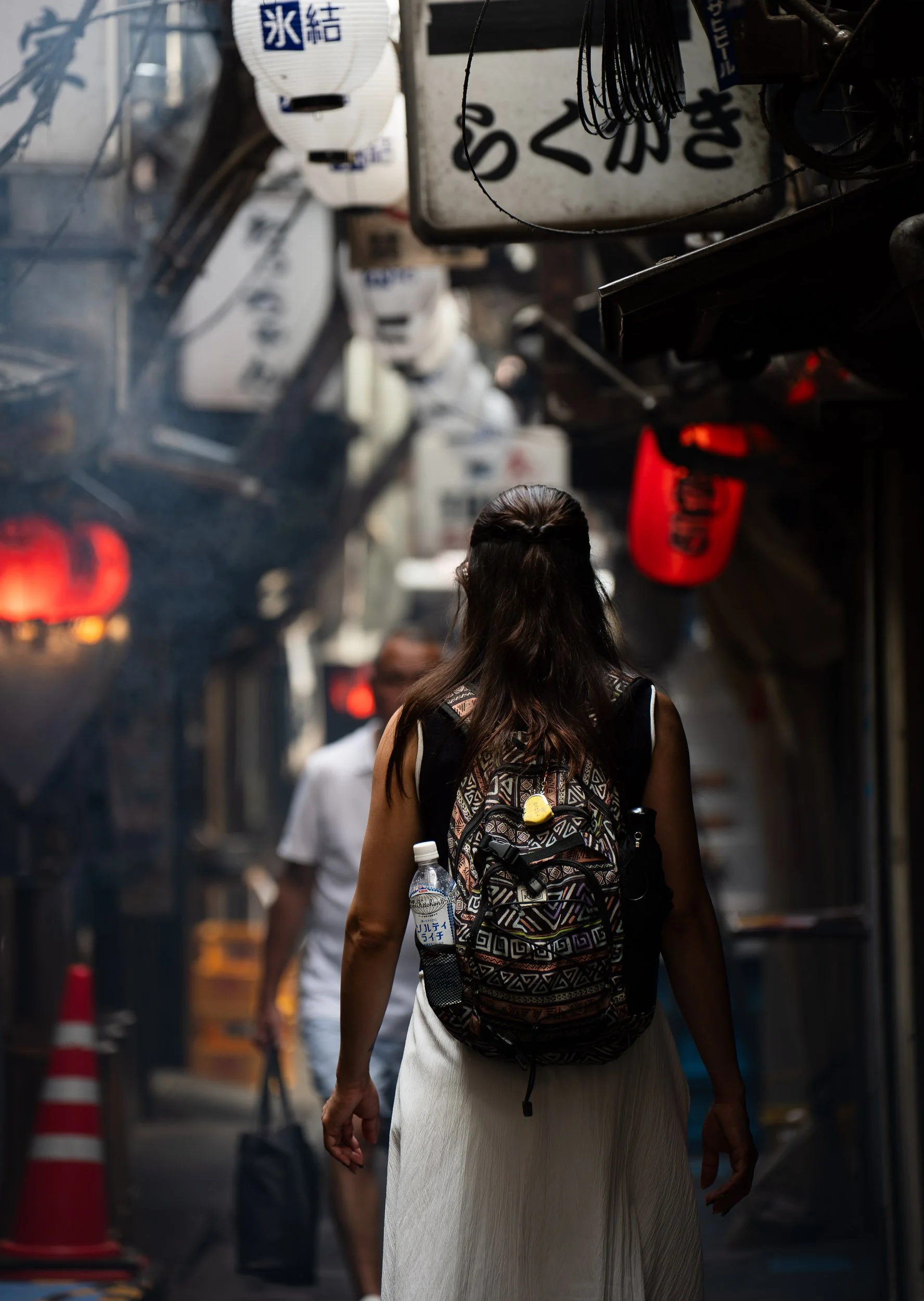 A woman with long dark hair, wearing a patterned backpack and a white skirt, walking through a busy street in Japan. The street has hanging lanterns with Japanese writing, signs, and red lanterns, with other pedestrians in the background.