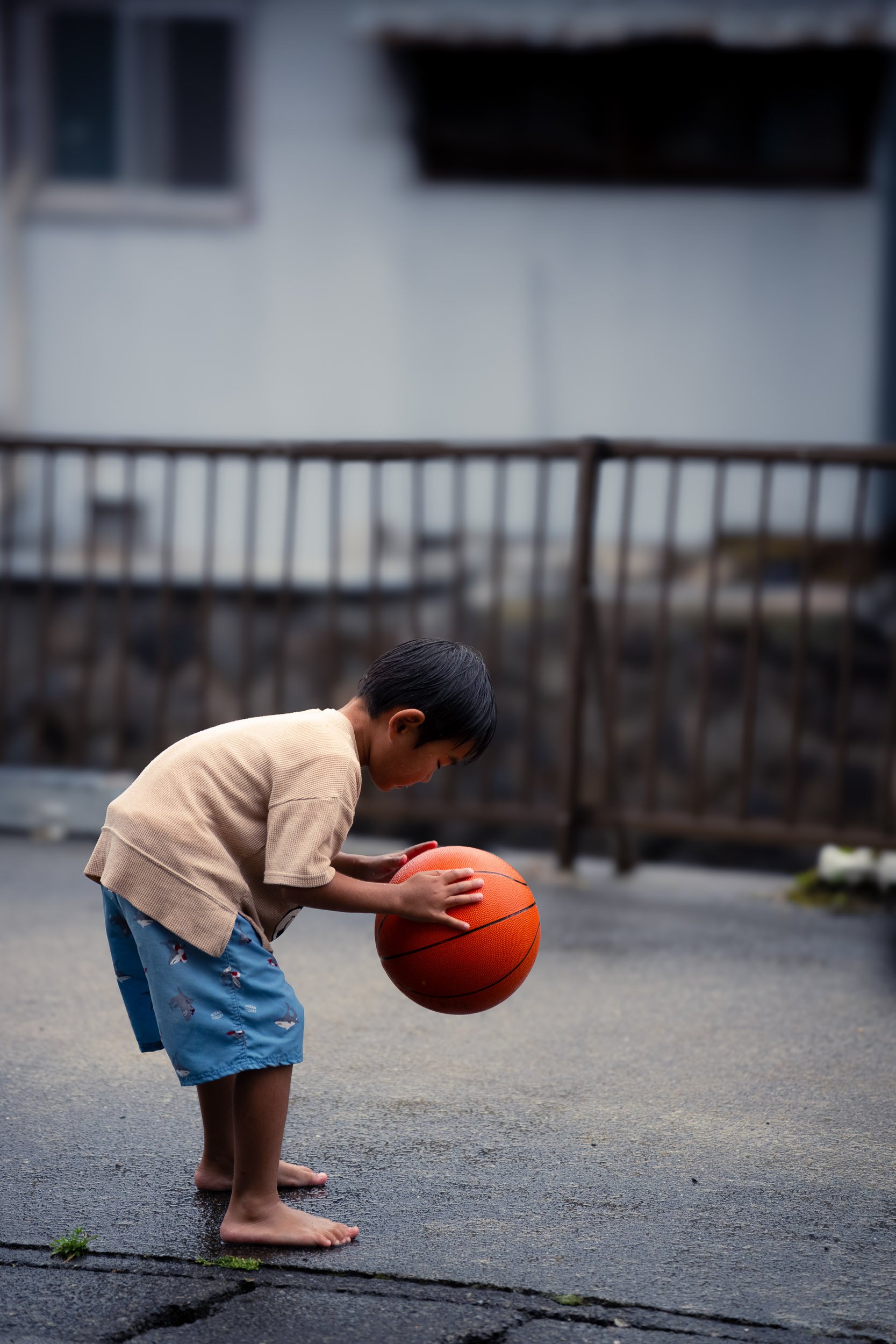 A young boy with short dark hair, barefoot, wearing a beige sweatshirt and blue shorts with colorful dinosaurs, is holding and looking down at an orange basketball on an outdoor asphalt surface, with a blurred background of a fence and building.