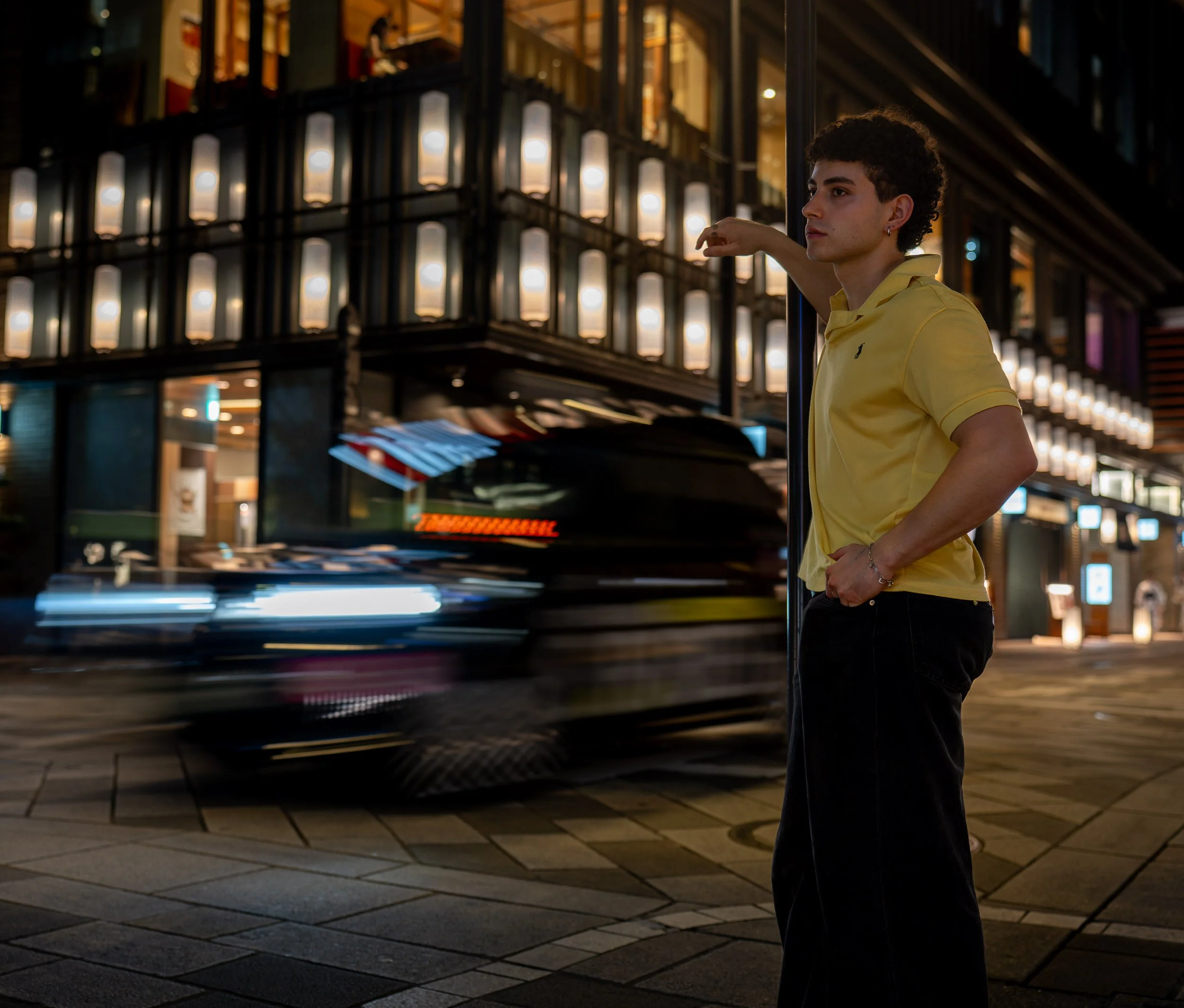 A young man in a yellow polo shirt stands at a city street corner at night, leaning against a lamp post with his right arm. Behind him, blurred moving cars and illuminated buildings are visible.