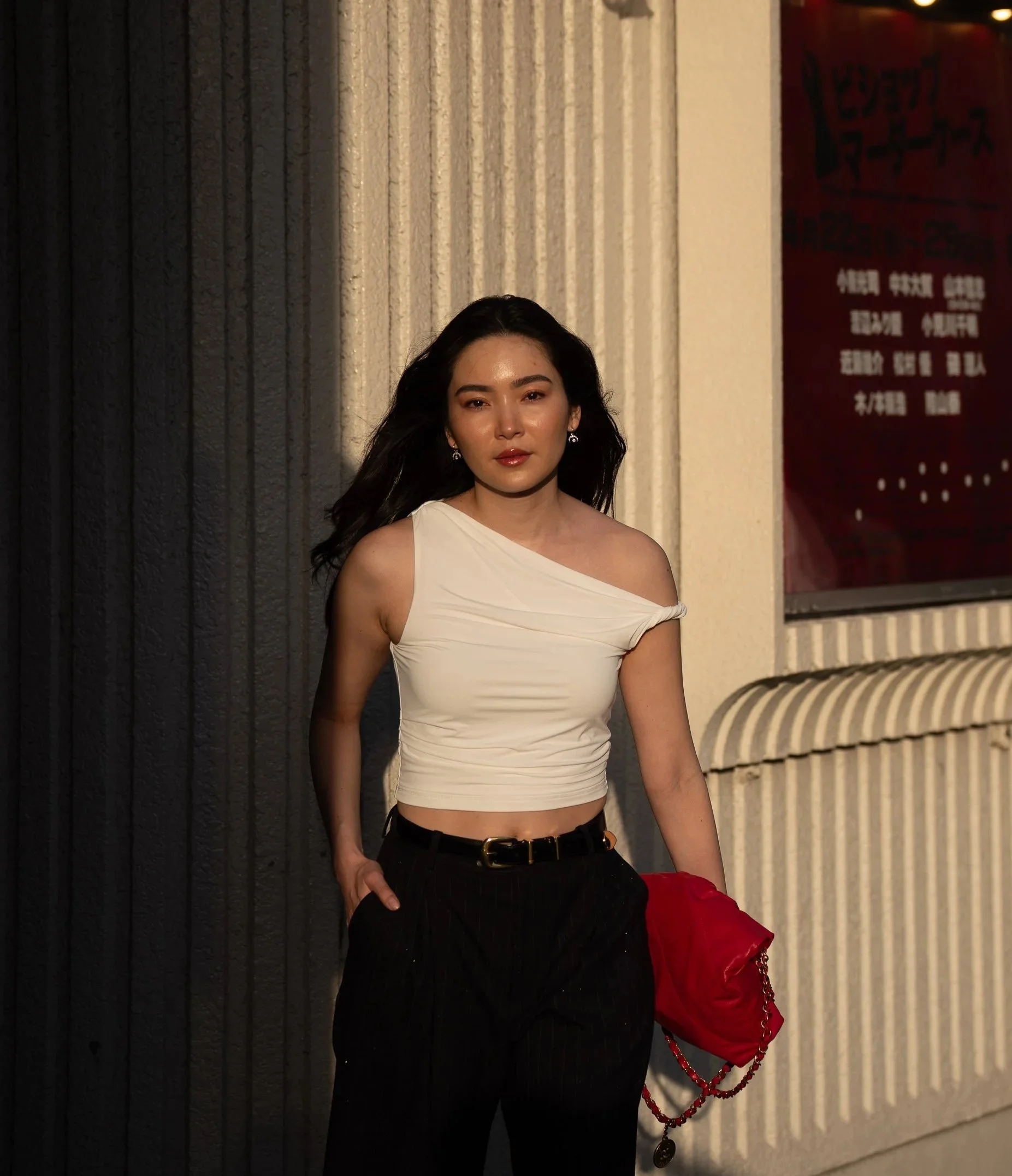 A woman with long dark hair, wearing a white off-the-shoulder top and black pants, standing outdoors at night against a textured wall, holding a red purse with chain details.