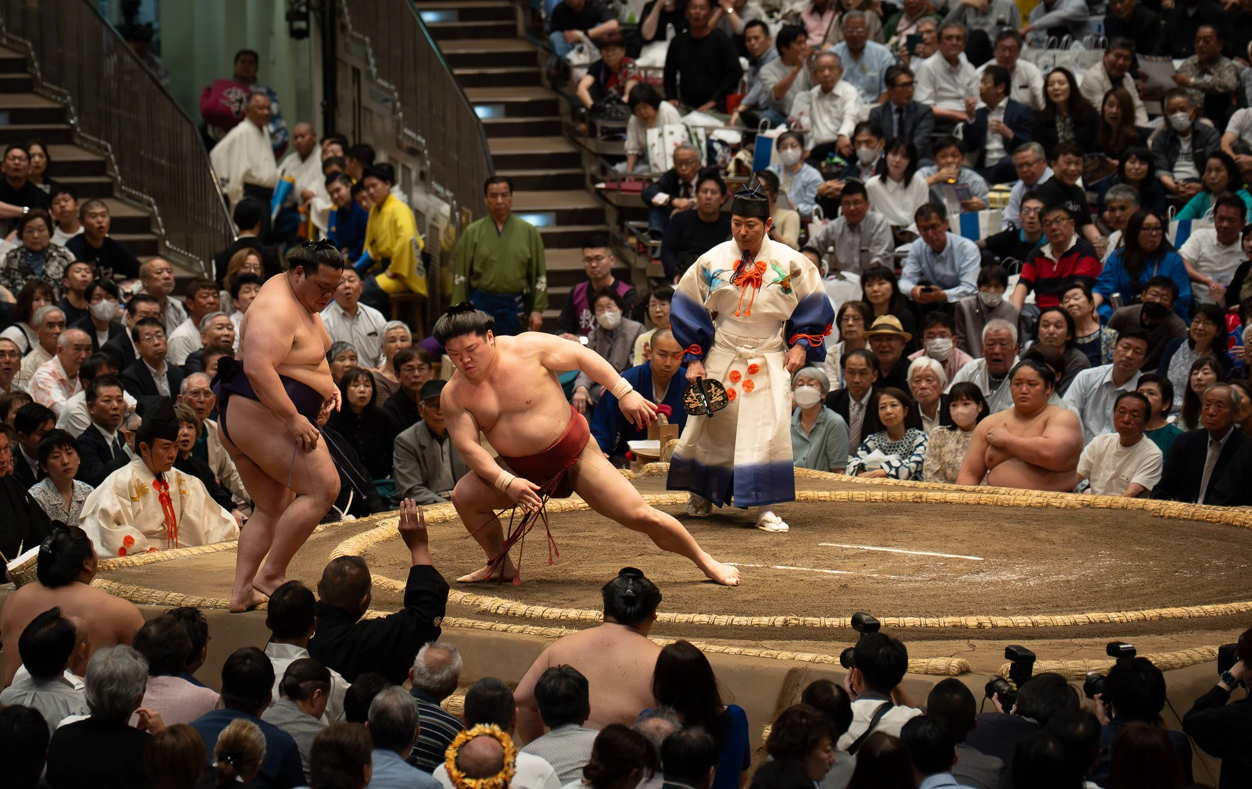 A sumo wrestling match in an arena with a large audience. Two sumo wrestlers are in the ring, engaged in combat, with one wrestler on the ground. A referee dressed in traditional attire is observing the match, surrounded by spectators.