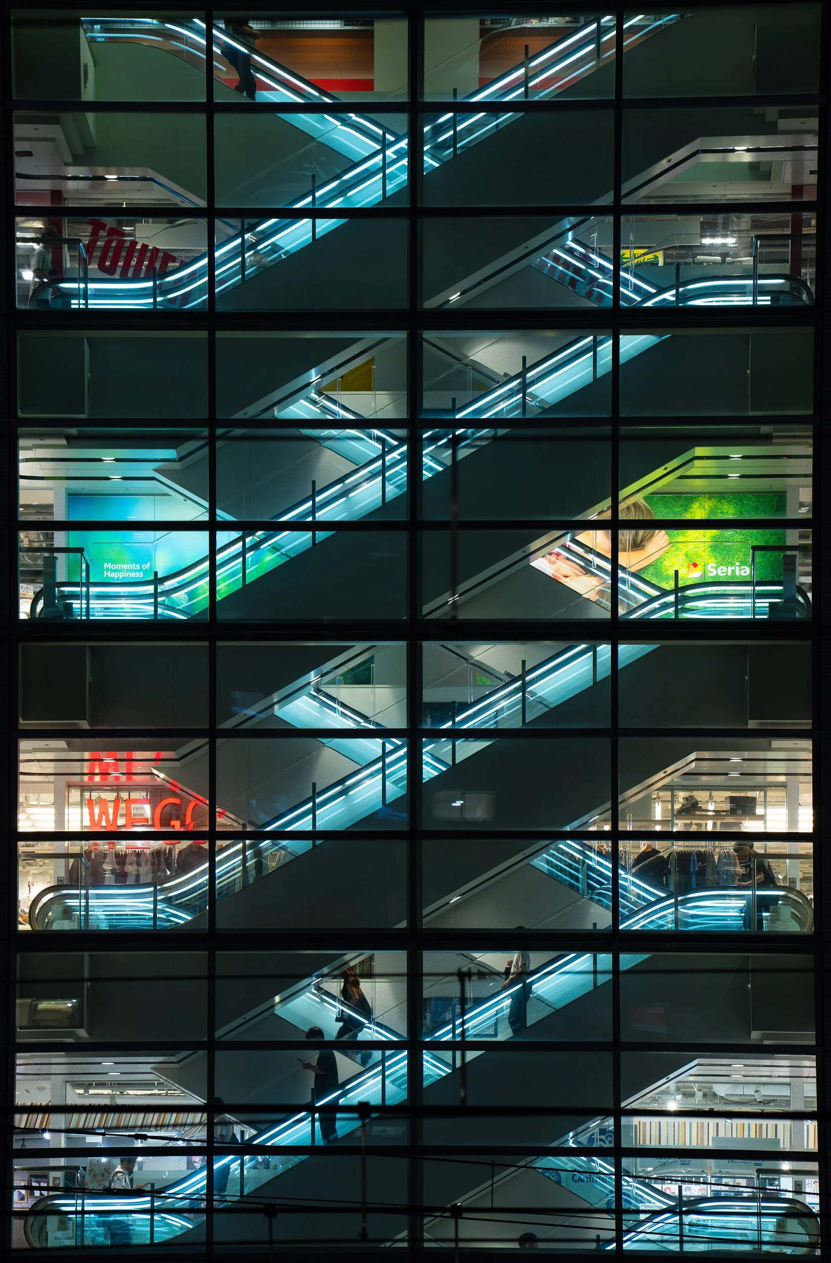 View of multiple escalators inside a modern shopping mall or commercial building, seen through large glass windows with black grid framing, illuminated with neon-like lighting.