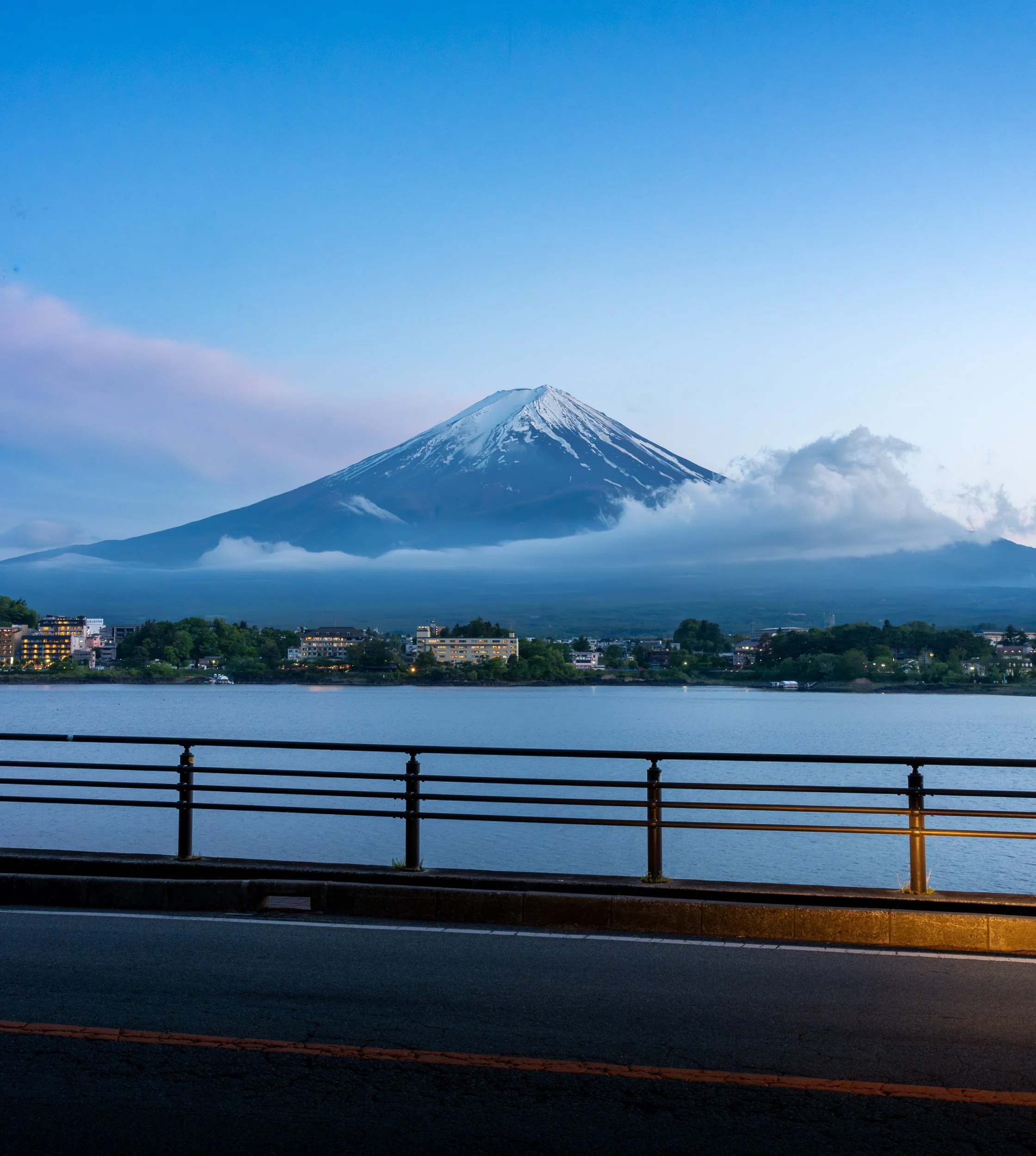 Mount Fuji with snow-capped peak and clouds around its base, viewed from a lakeside promenade with a metal railing and road in the foreground, under a partly cloudy sky.