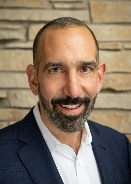 Close-up of a smiling man with short hair, a beard, wearing a dark blazer and white dress shirt, standing in front of a brick wall.