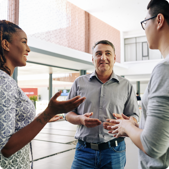 Three people having a conversation indoors, smiling and gesturing, with a modern building background.