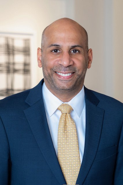 portrait of a man in a navy suit and yellow tie smiling in an indoor setting.