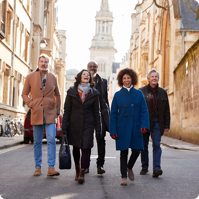 A diverse group of five friends walking together on a city street during daytime, smiling and laughing, with historic buildings and a church tower in the background.