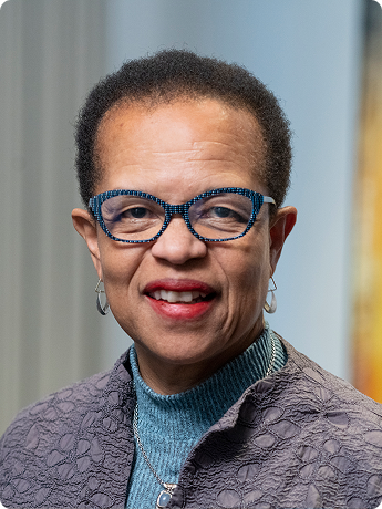 A woman with short curly hair wearing glasses, earrings, a blue turtleneck, and a purple textured jacket.