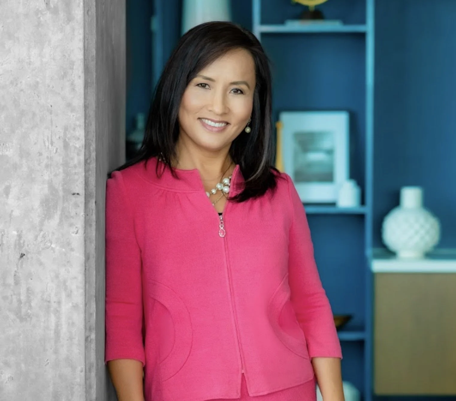 A woman with shoulder-length dark hair, wearing a bright pink jacket and pearl jewelry, smiling and leaning against a concrete wall in a modern room with blue shelving and decorative objects.