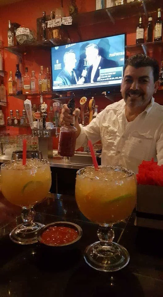 A bartender behind a bar counter with two large cocktail drinks with red straws and lime wedges, holding a bottle of liquor. A man in a white shirt is smiling, and a TV screen shows two people in an interview or discussion in the background.