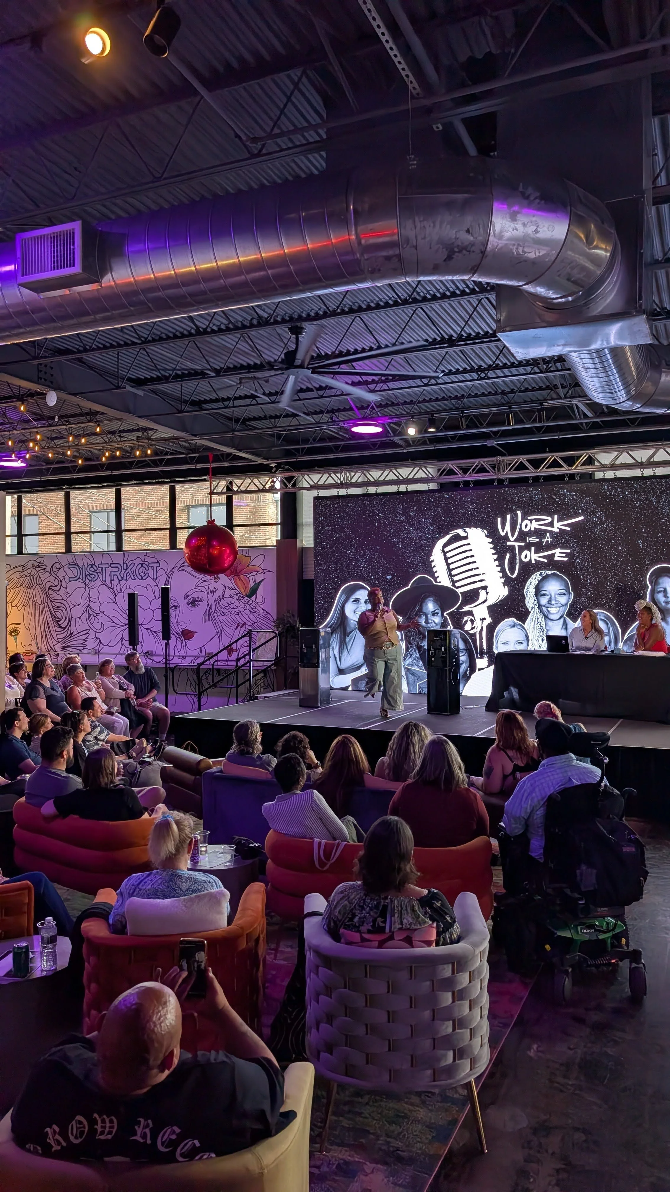 An indoor event with a stage featuring a large black and white screen displaying the words 'Work is a Joke' with a microphone graphic. Audience members are seated in colorful chairs, watching a person on stage speaking into a microphone. The room has industrial-style decor with visible ductwork and purple accent lighting.