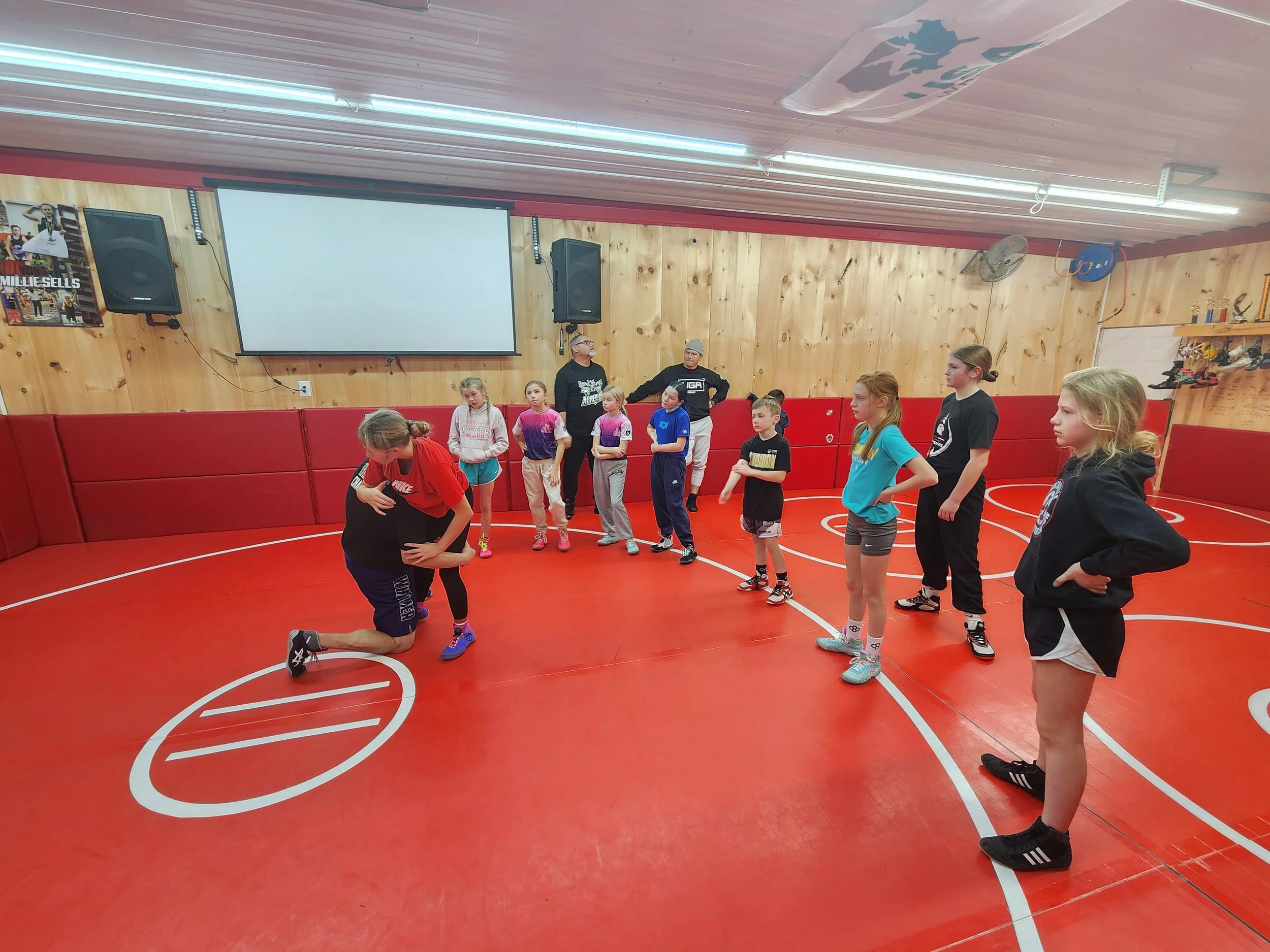 A group of children and two instructors standing on a red indoor sports court, with a large white projection screen on a wooden-paneled wall behind them, in a gym or sports facility.