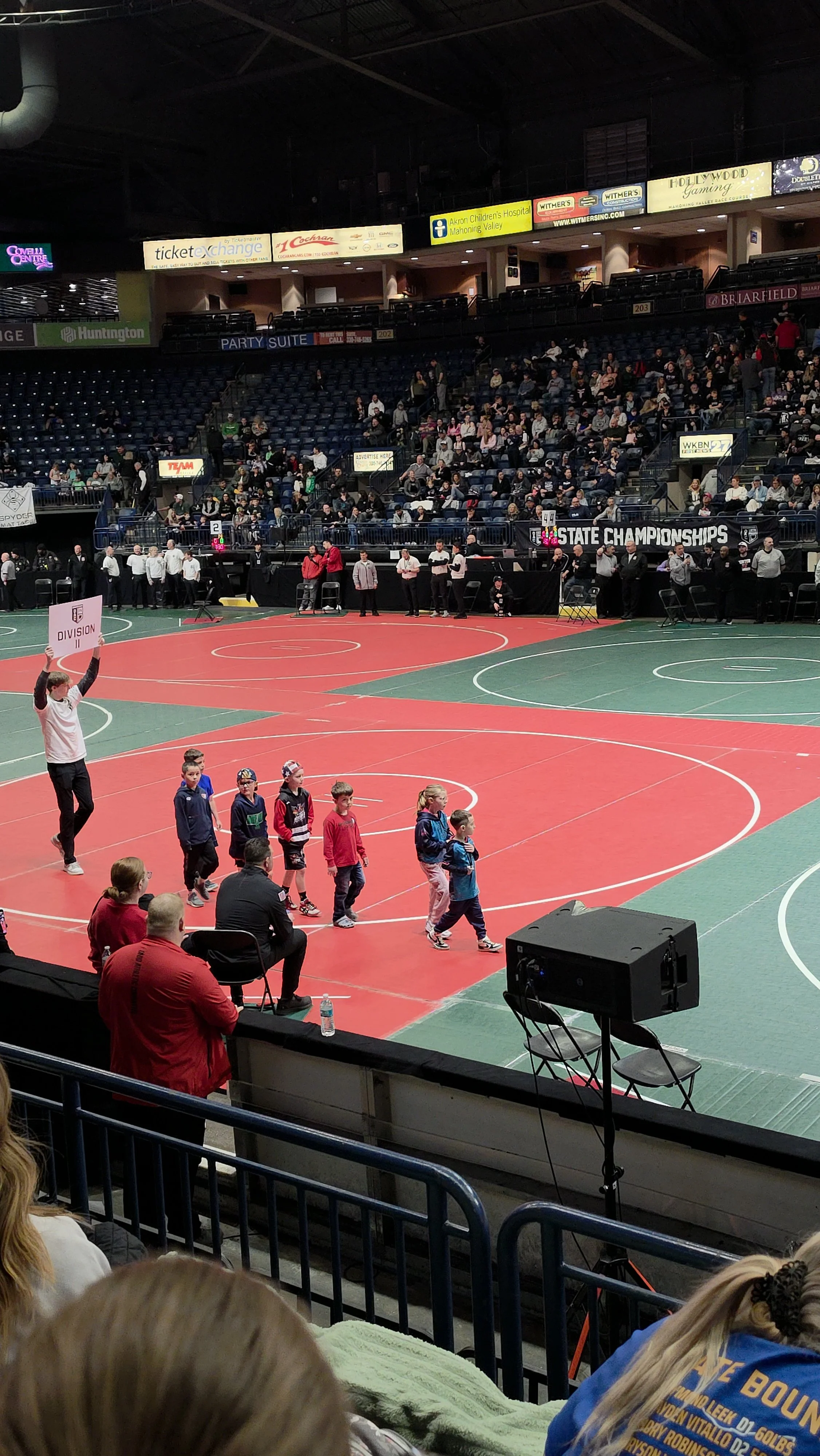 Children walking on the edge of an indoor sports court during a wrestling event with spectators watching from the stands.