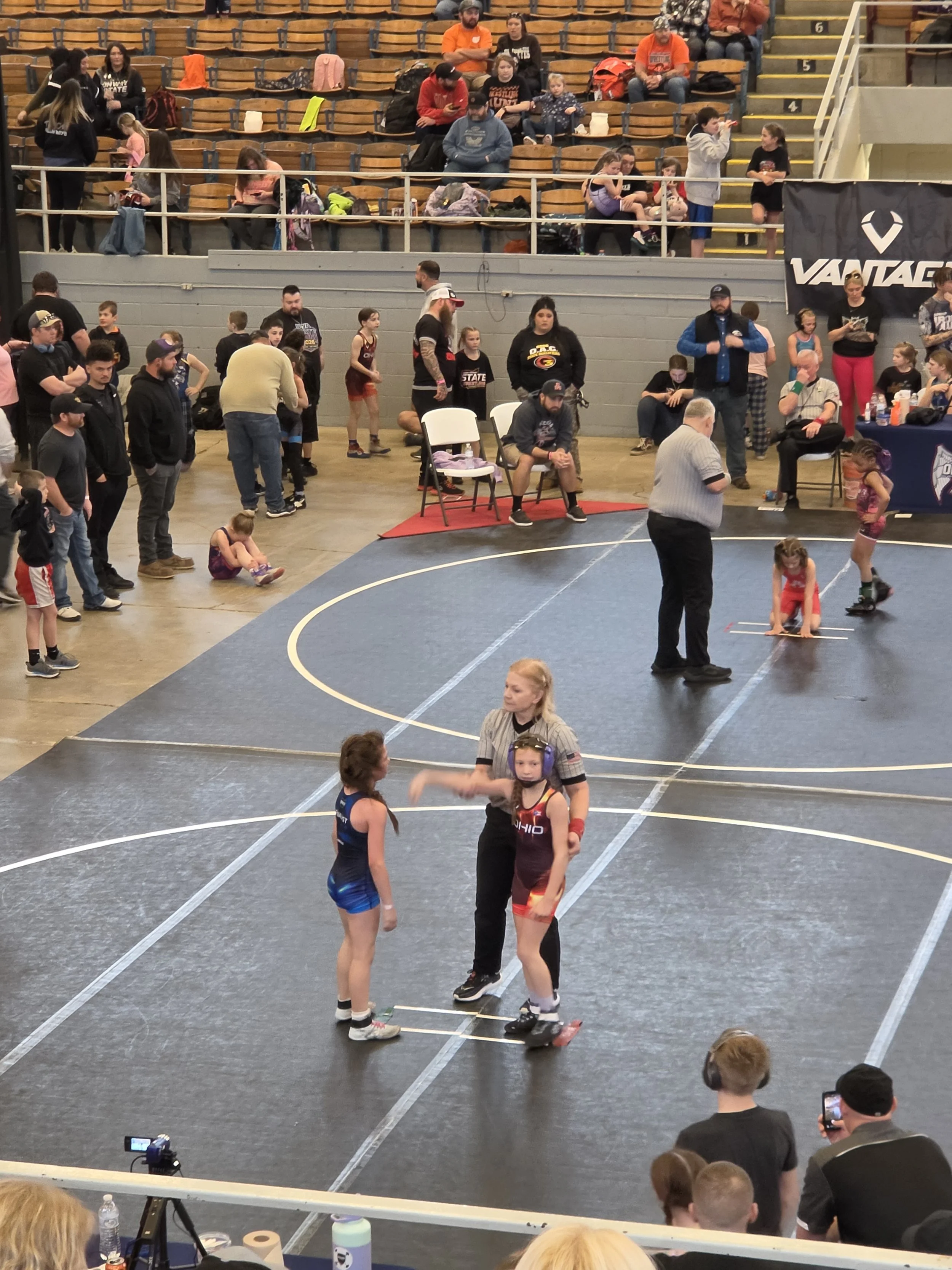 A wrestling match taking place in an indoor gymnasium with young athletes. Two girls are standing on the mat, seemingly preparing for a match or coaching. Spectators, including children, sit in the bleachers and around the mat, watching and taking photos. Some people are seated while others are standing or walking around.