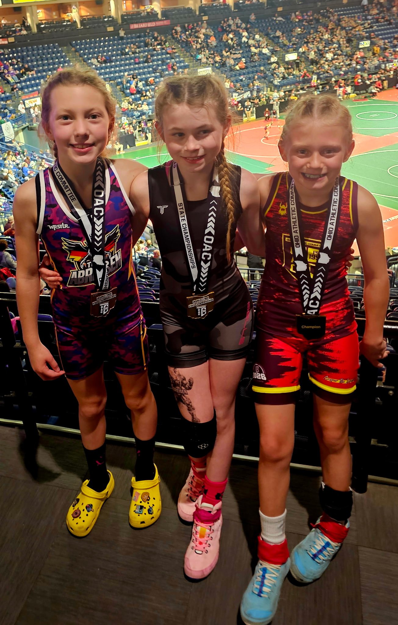 Three young girls standing side by side in a sports arena, wearing athletic uniforms and medals around their necks. They are smiling, and the background shows a basketball court with spectators in the stands.