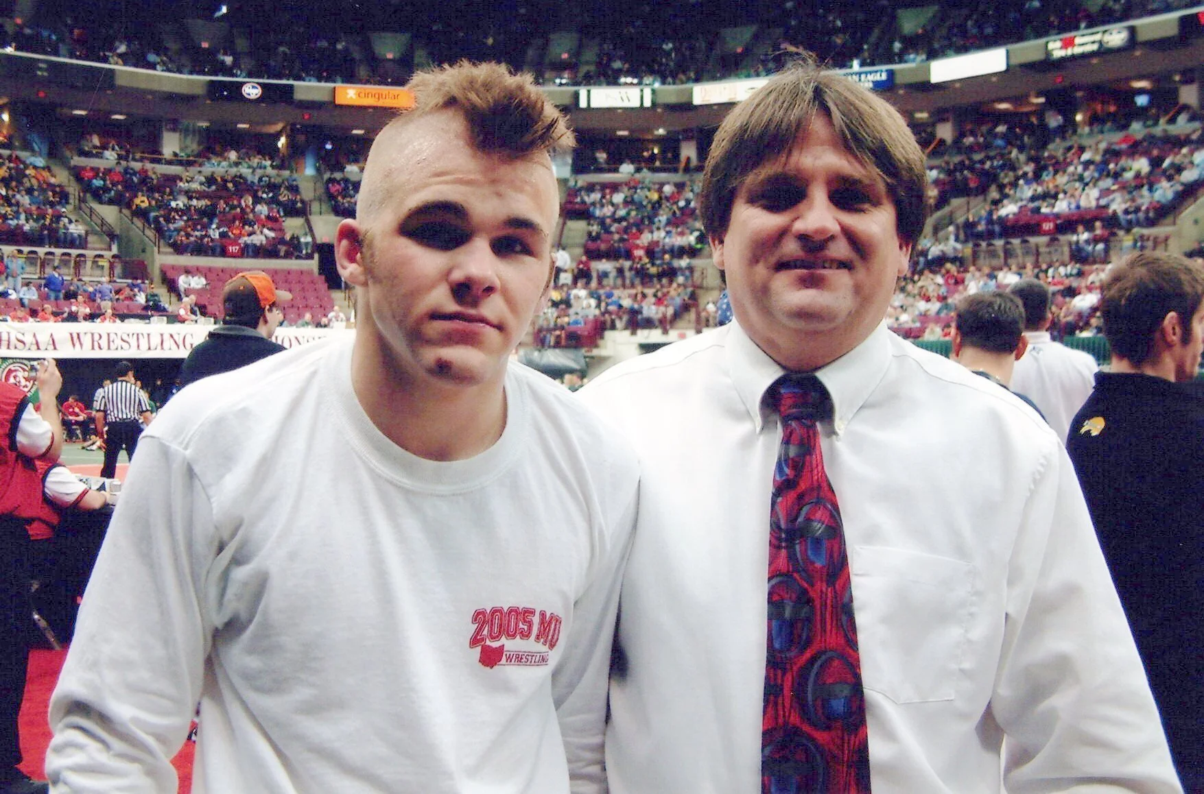 Two men standing side by side, in an arena with a crowd in the background. The man on the left has a mohawk hairstyle and is wearing a white t-shirt. The man on the right has straight brown hair, is smiling, and is wearing a white shirt with a colorful tie.