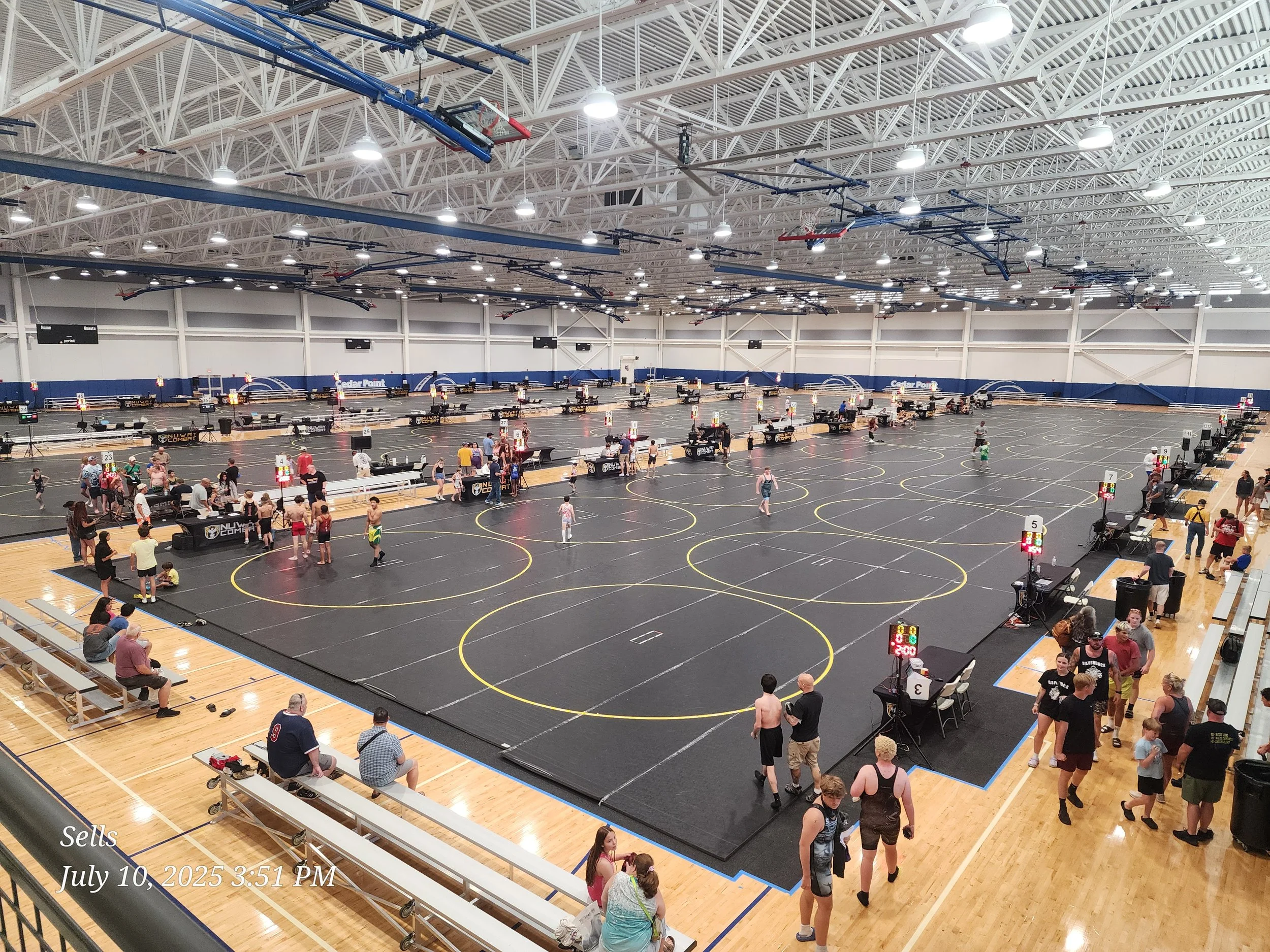 An indoor sports facility with multiple 3-on-3 basketball courts. People are standing, sitting, and walking around the courts, some with bags and equipment. The ceiling has numerous lights and hanging equipment, and there are scoreboards on the walls. The floor is wooden, and the space is spacious and well-lit.