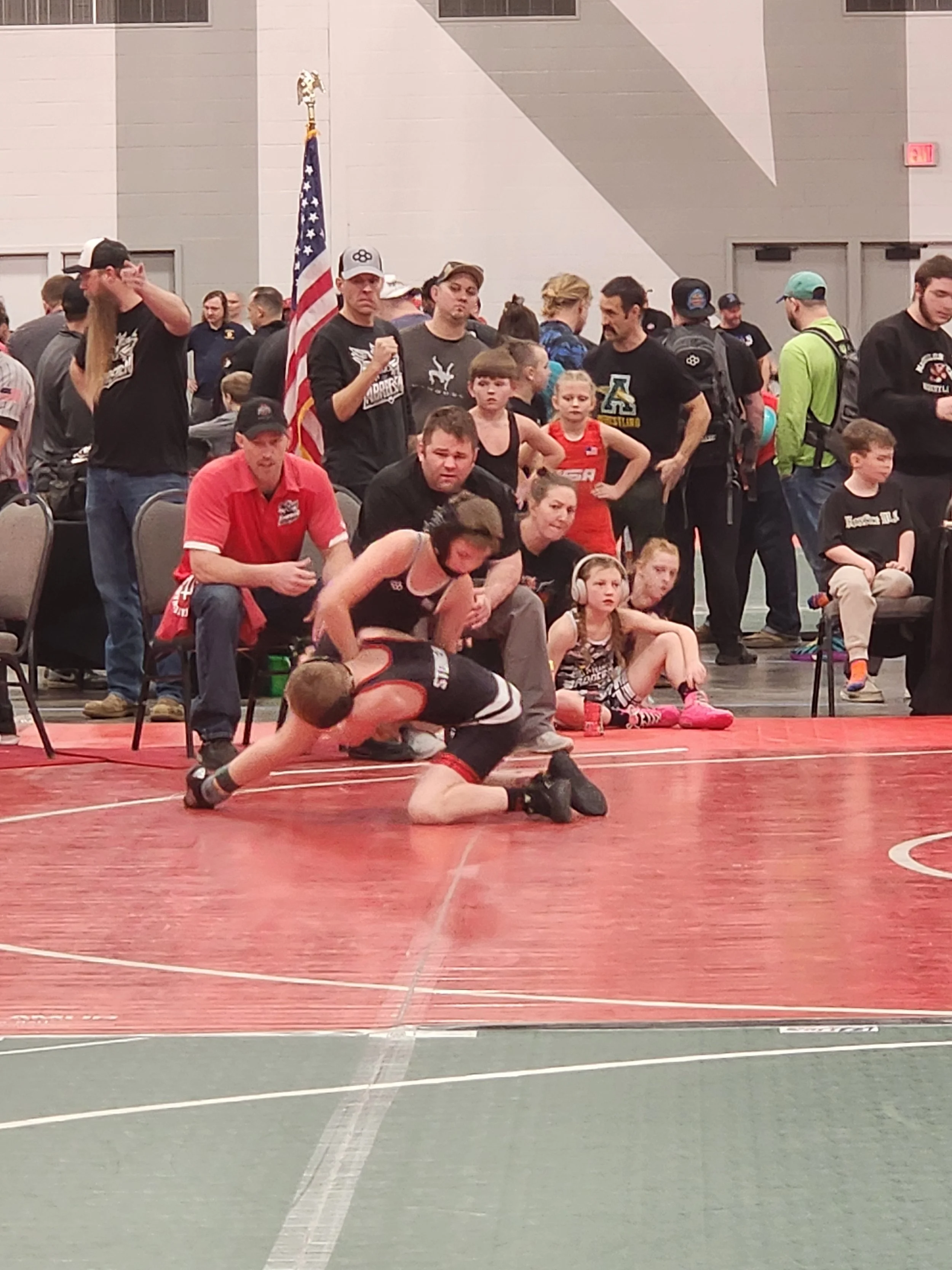 Young male wrestler in black and white singlet on a wrestling mat, being assisted by a woman, with crowd and people watching in the background.
