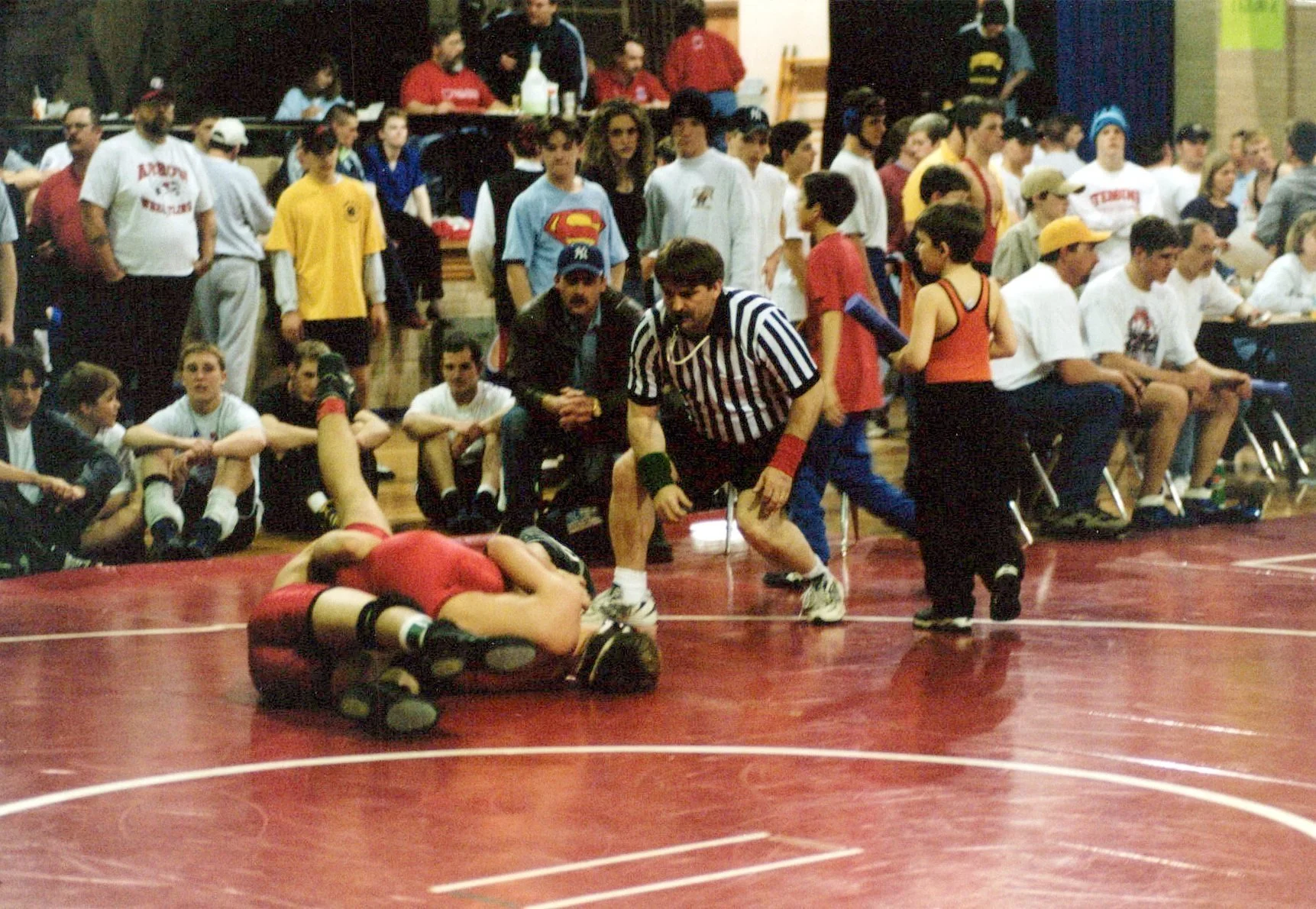 A wrestling match is taking place on a red mat with children and teenagers sitting and standing around watching. One wrestler is lying on the mat, while another wrestler kneels nearby. Two referees monitor the match, one in the foreground wearing a black and white striped shirt. Spectators are seated in chairs, some holding papers or clipboards, and others are standing in the background.