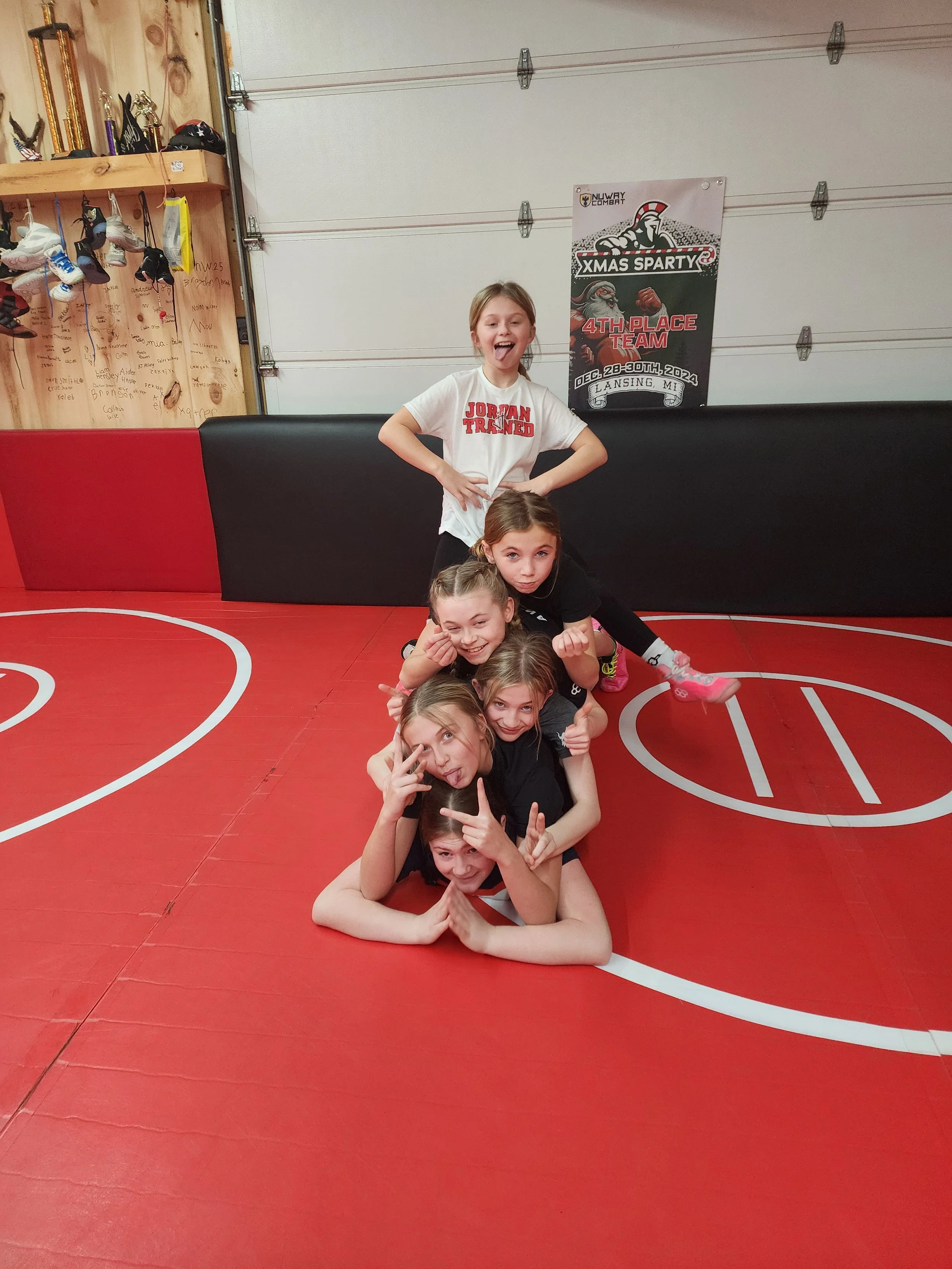 A group of seven young girls posing on a red basketball court, making playful gestures. Behind them is a wall with a poster and a wooden wall with shoes hanging.