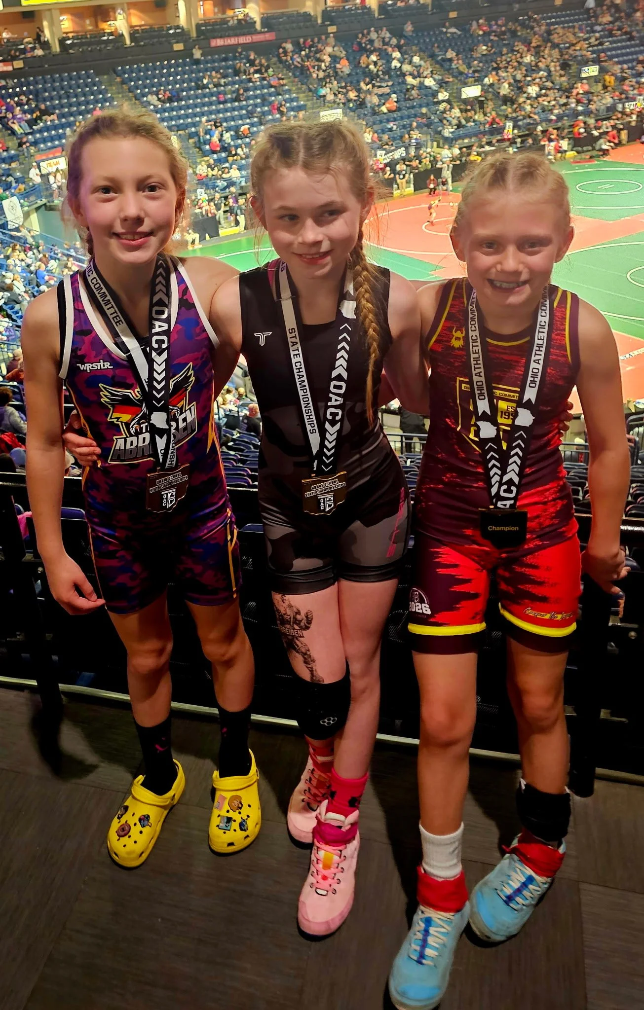 Three young girls in athletic outfits and medals around their necks at a basketball game in an arena. The girl on the left is wearing a colorful uniform with a logo and yellow Crocs, the girl in the middle is dressed in black and pink with pink shoes, and the girl on the right is in red and black with blue shoes.