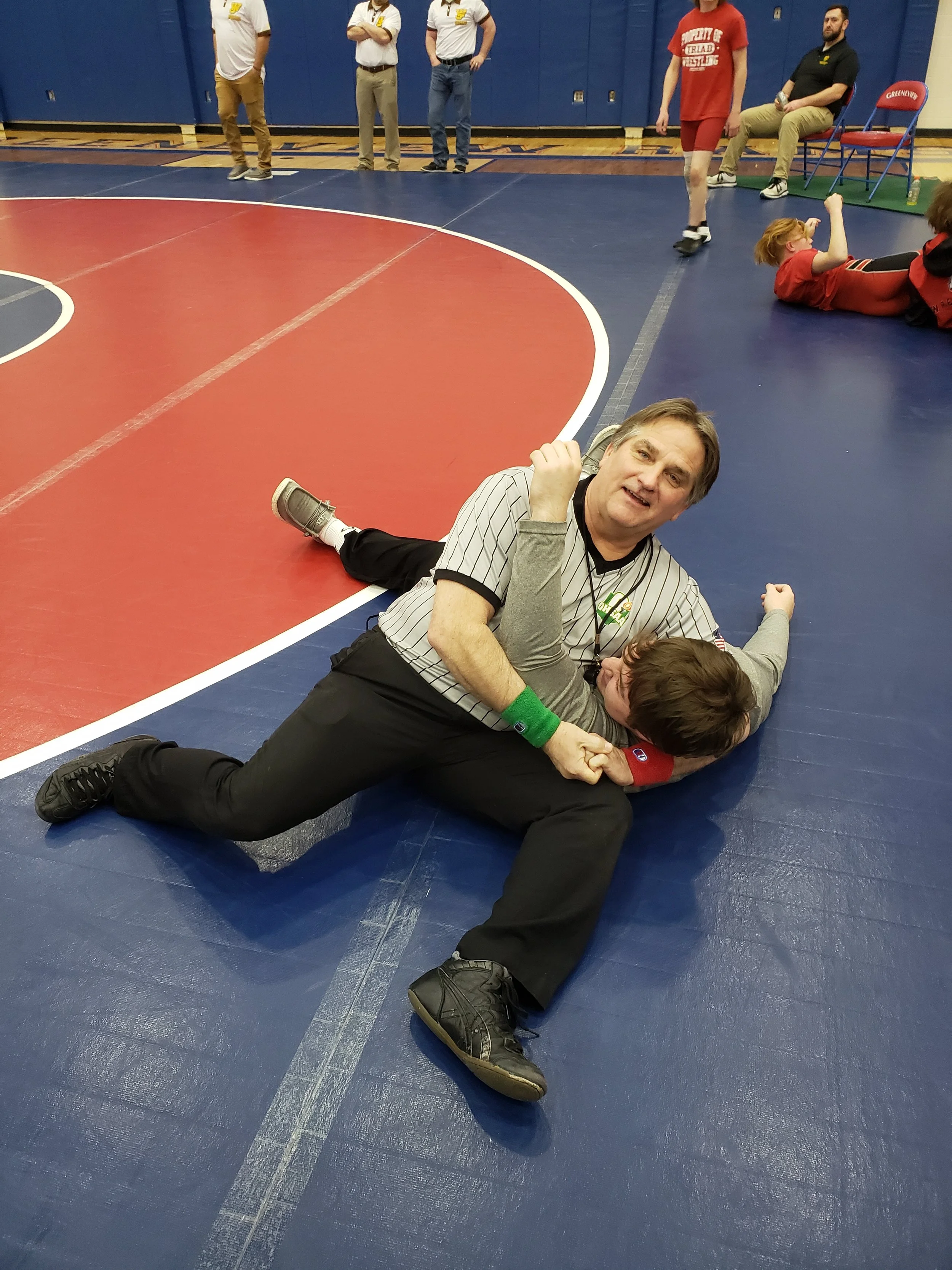 A wrestling match with a referee on the mat pinning a young wrestler while an adult looks on. Other young wrestlers and adults are in the background in a gymnasium.