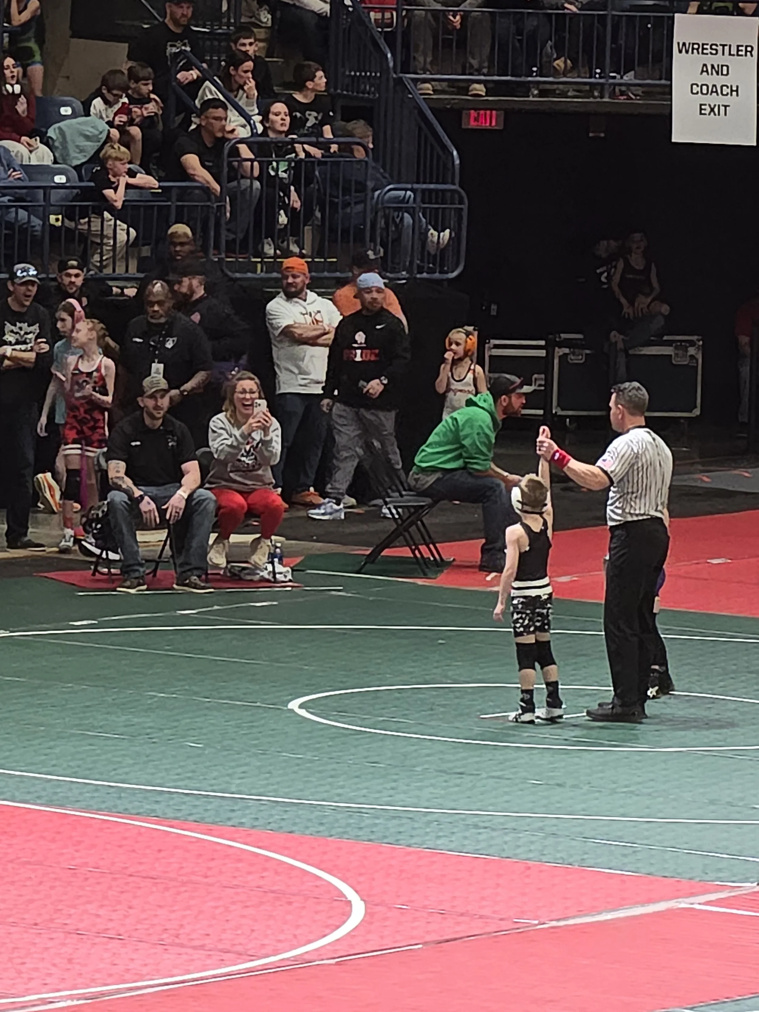 A wrestling match is taking place in an indoor sports arena. Two young wrestlers are in the ring, facing each other, with a referee standing nearby. Spectators are seated around the ring, watching the match.