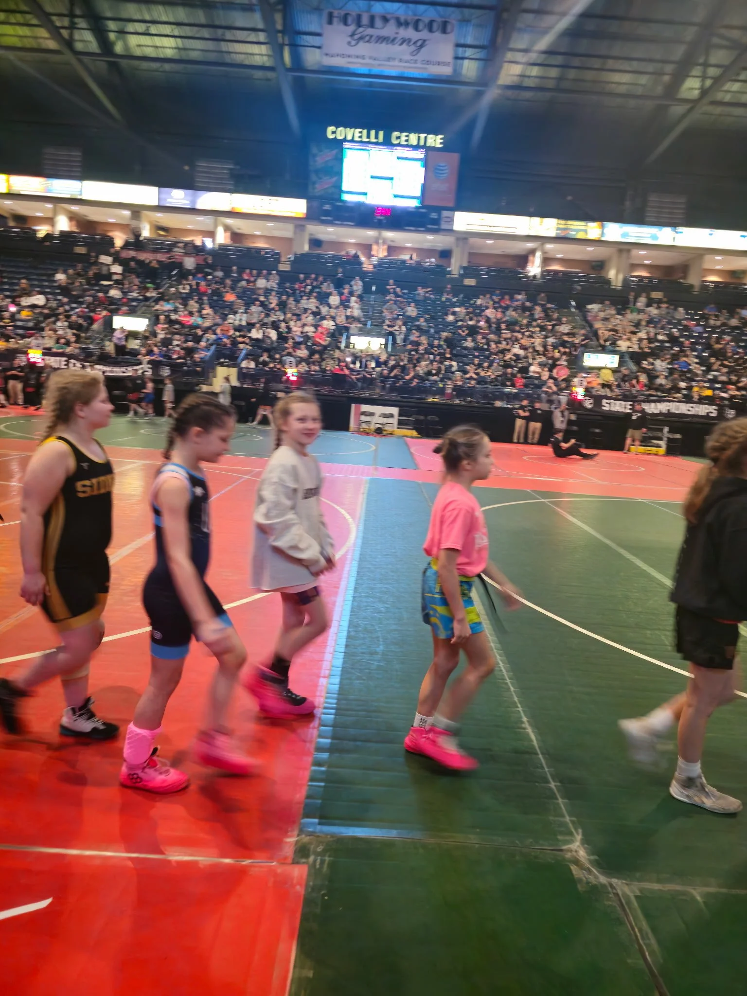 Indoor sports arena with children walking in a line on a multicolored court, audience seated in the stands, scoreboard and signs visible above.