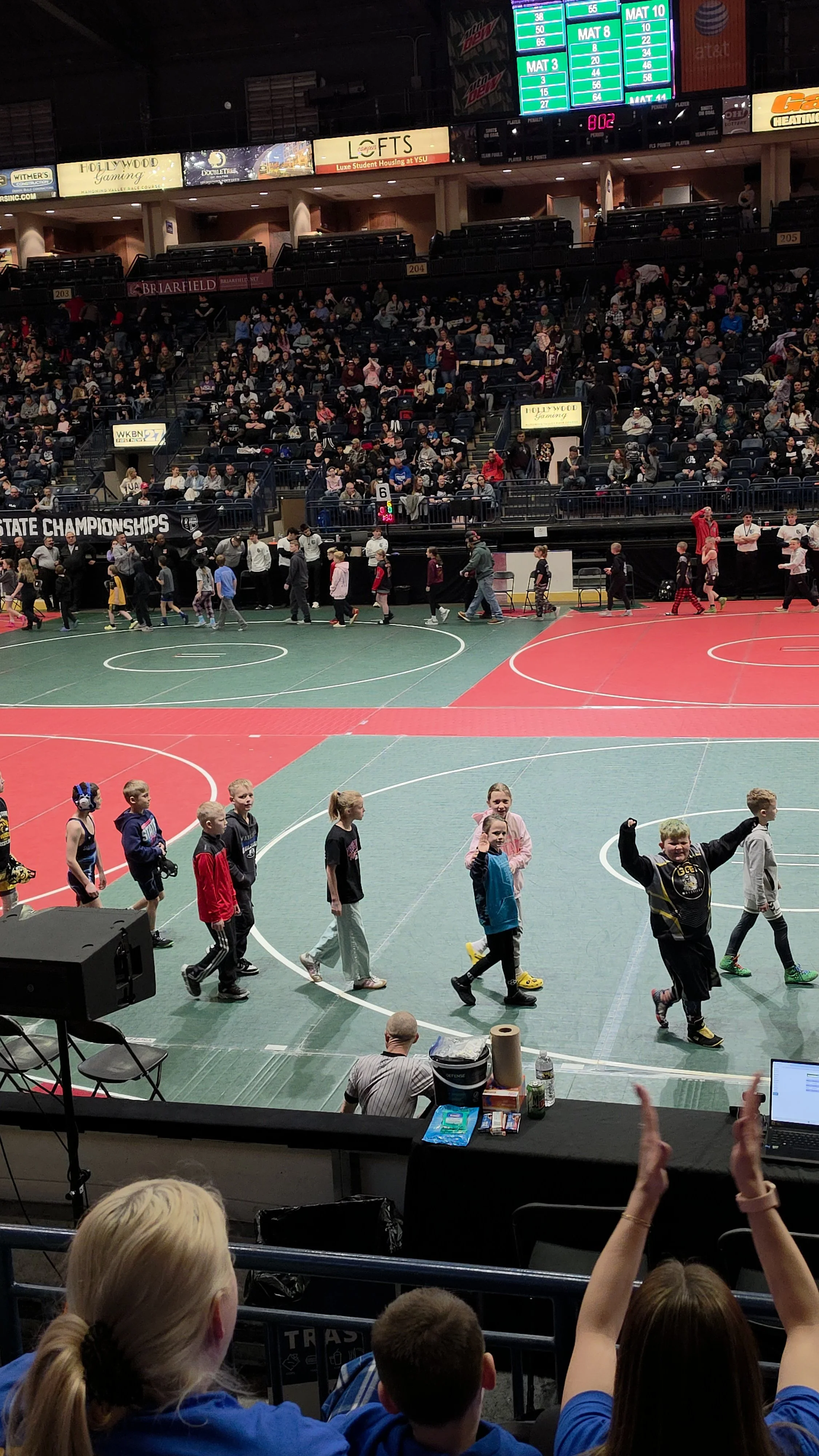 Children and young teens walking across an indoor gymnasium floor with a red and green mat, spectators in the bleachers, and scoreboard screens hanging above.