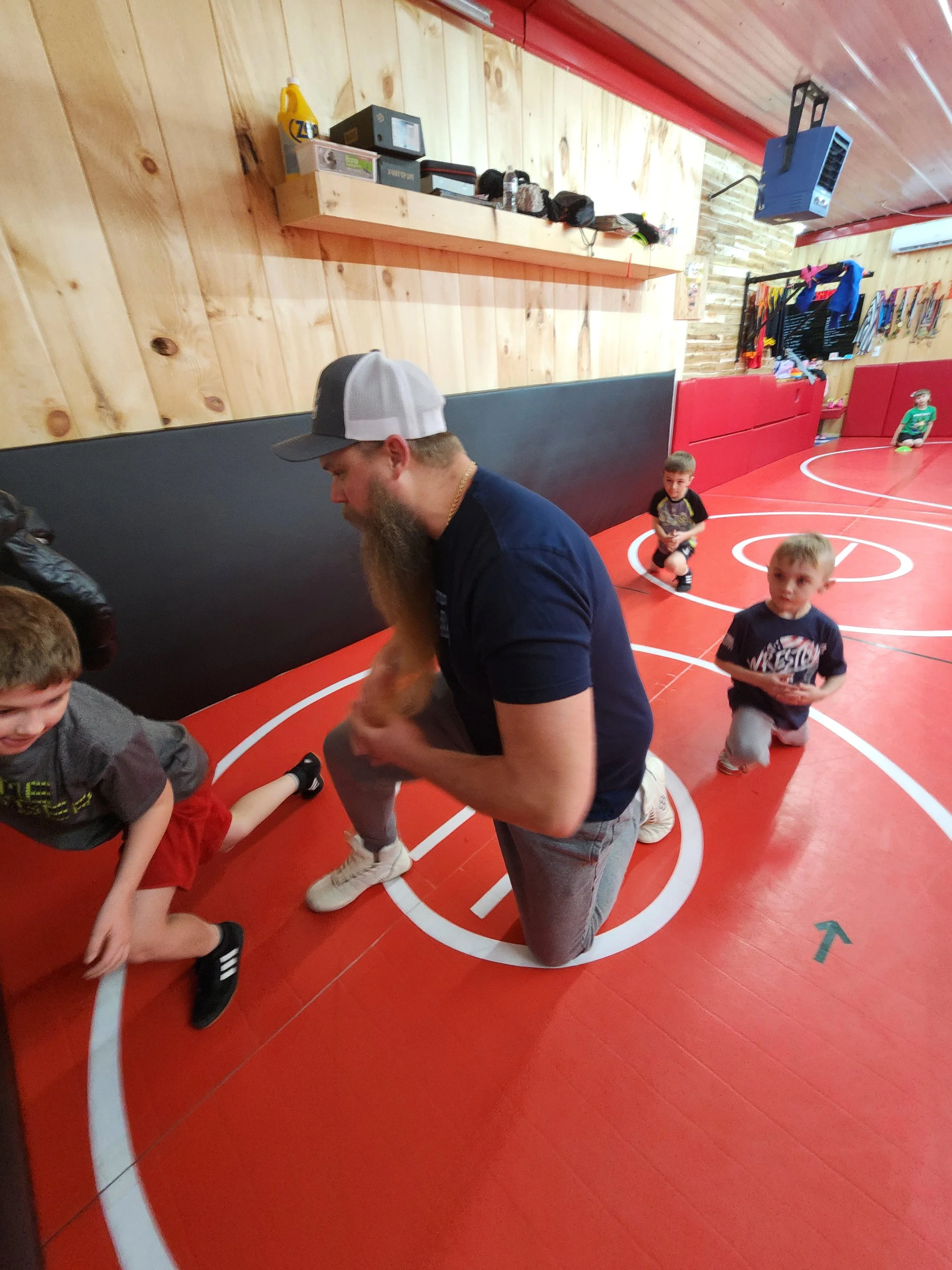 A man with a beard and cap kneeling on the floor of a basketball gym, engaging with children during a sports activity.