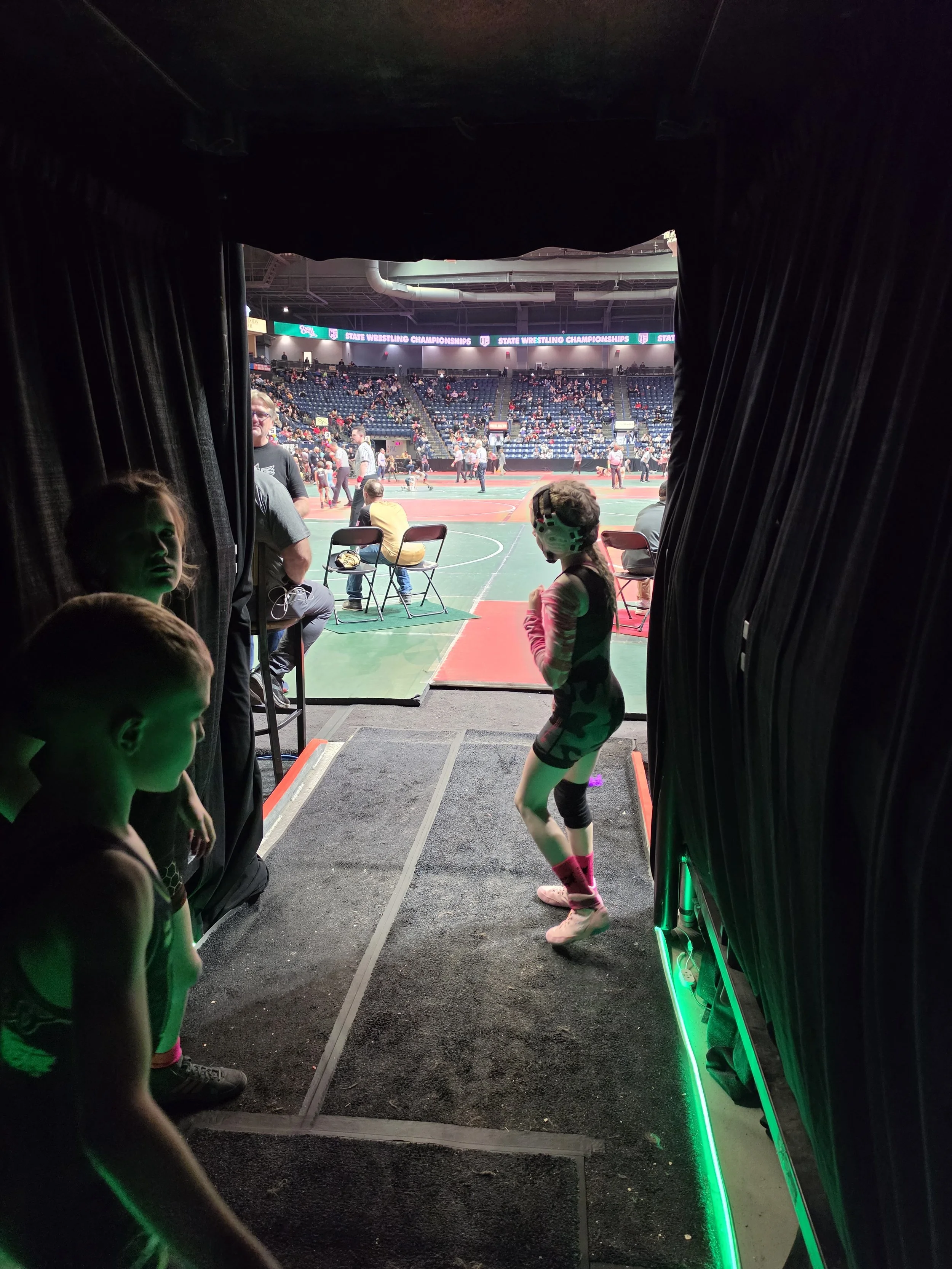 View from backstage of a wrestling event showing young wrestlers and people in the arena, with a sports arena and audience in the background.