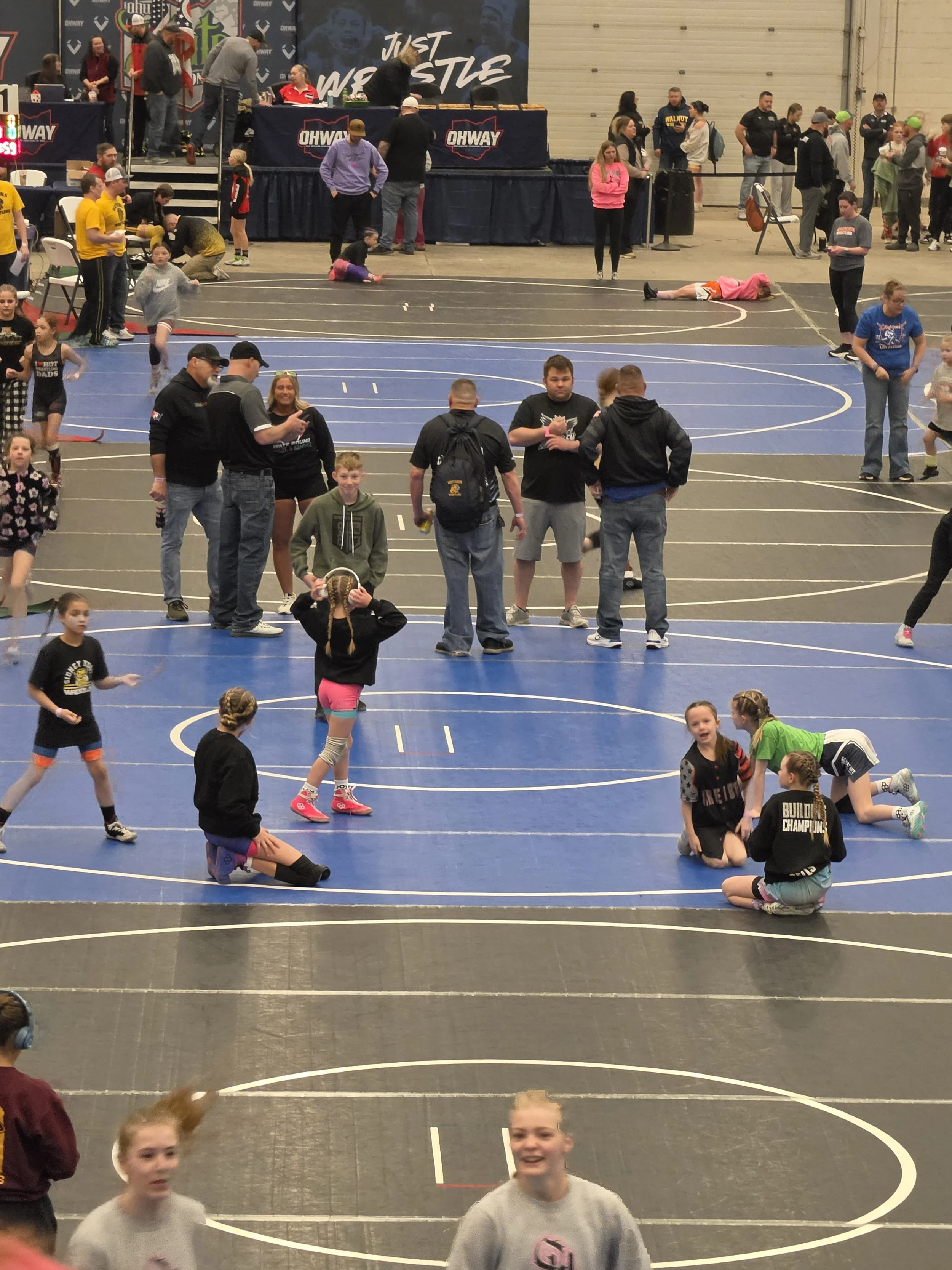 Children and adults inside a gymnasium participating in a sporting event or activity, with some kids running or playing and others waiting or walking around, and event banners and spectators in the background.