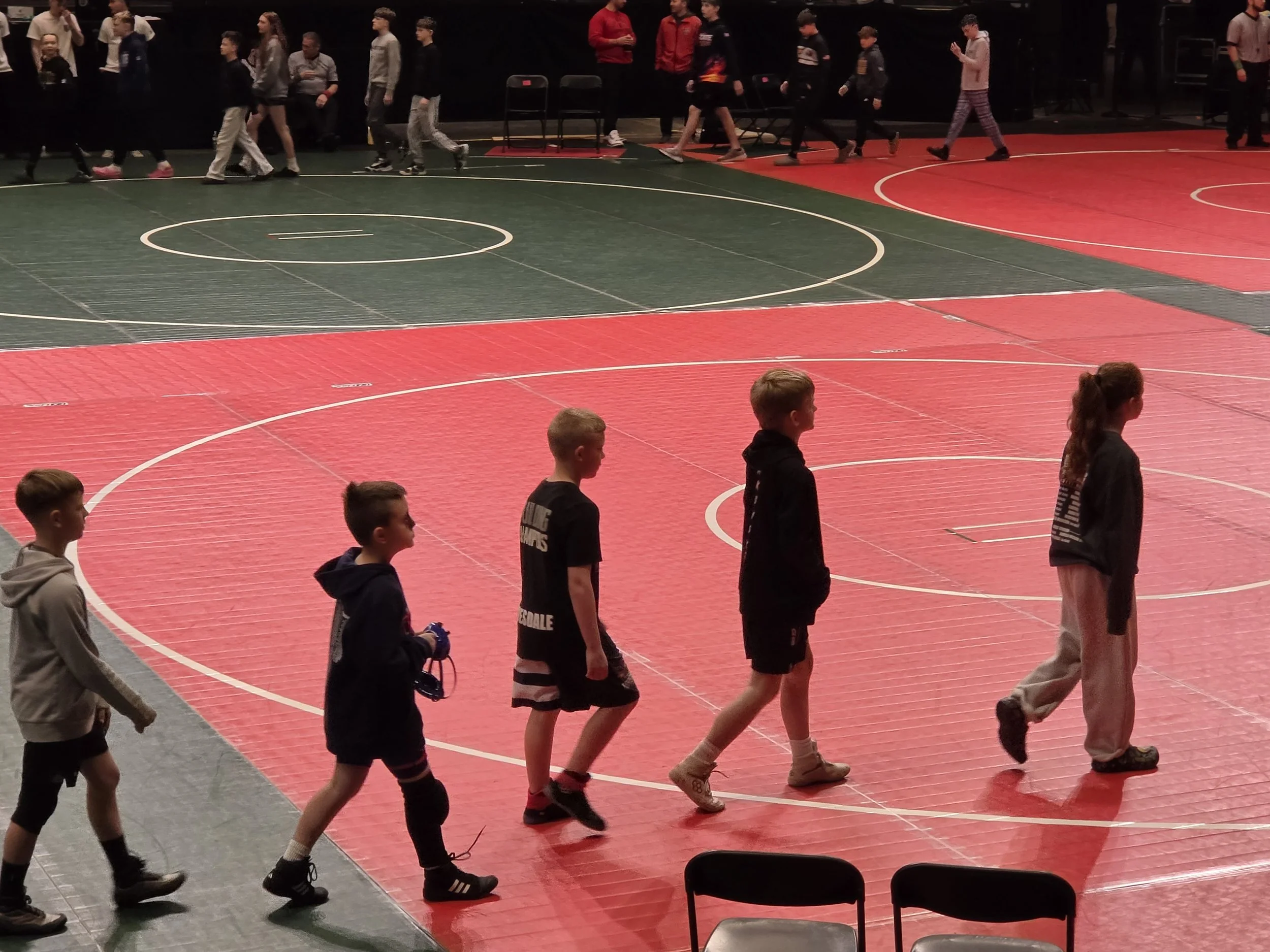Children walking in a line on a basketball court inside a gymnasium, with some adults sitting and standing in the background.
