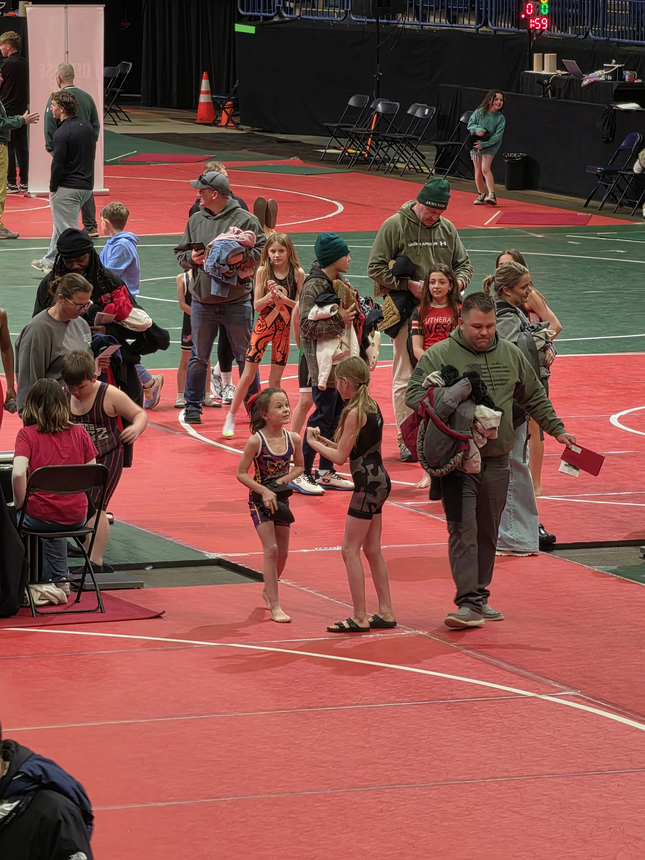 People gathered in an indoor gymnasium, some standing and some sitting, with two young girls in athletic wear talking on a red mat. There are chairs and equipment along the walls and a digital timer on the wall.