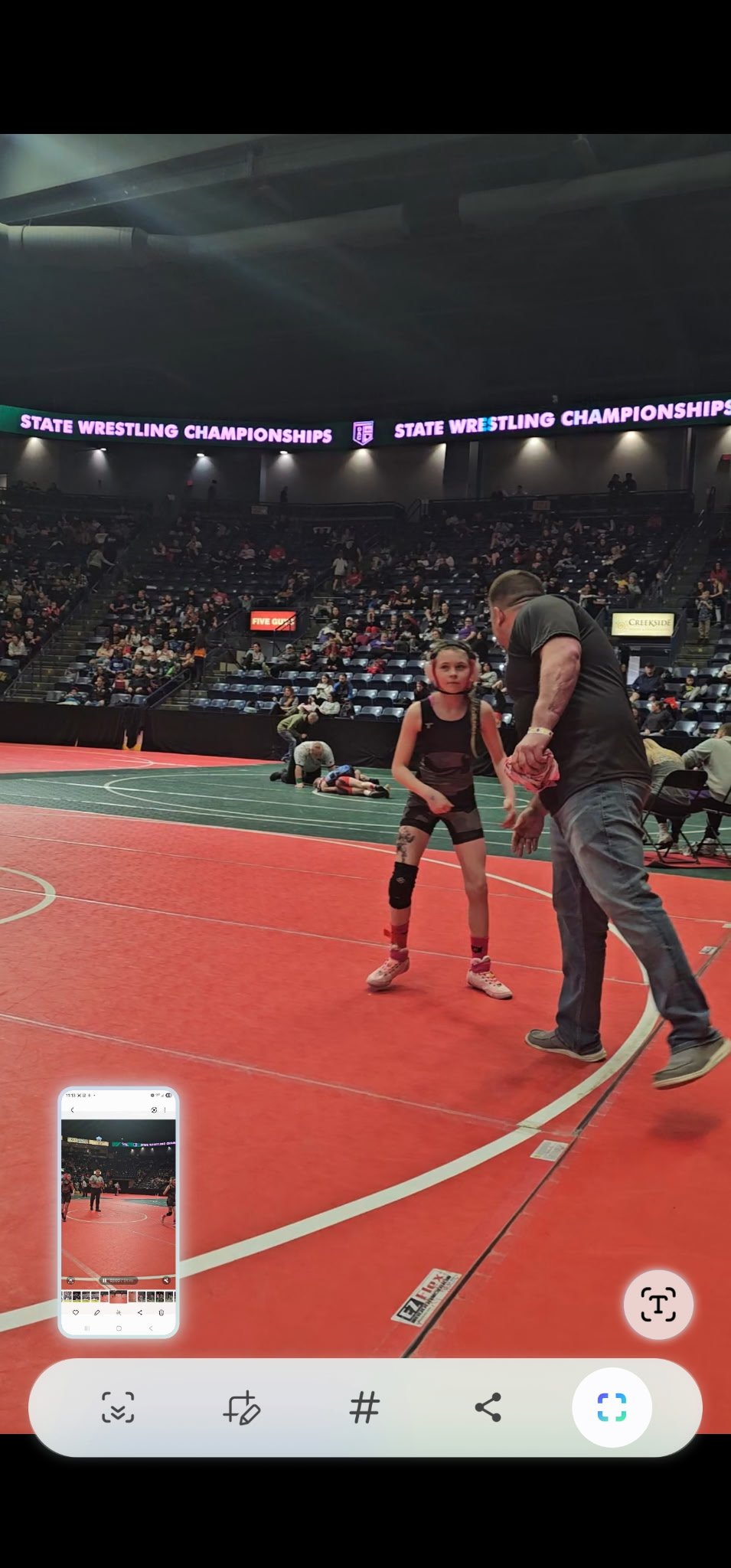 At the state wrestling championships, a young female wrestler is talking to her coach on the wrestling mat, while spectators are seated in the stands in the background.