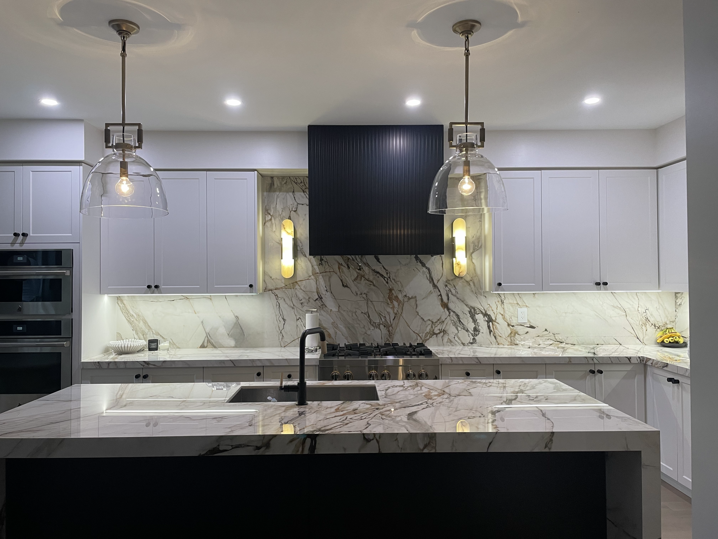 Modern kitchen with white cabinets, marble backsplash, and a marble countertop island. Pendant lights hanging from the ceiling and built-in oven visible on the left. Small decorative items on the counter.