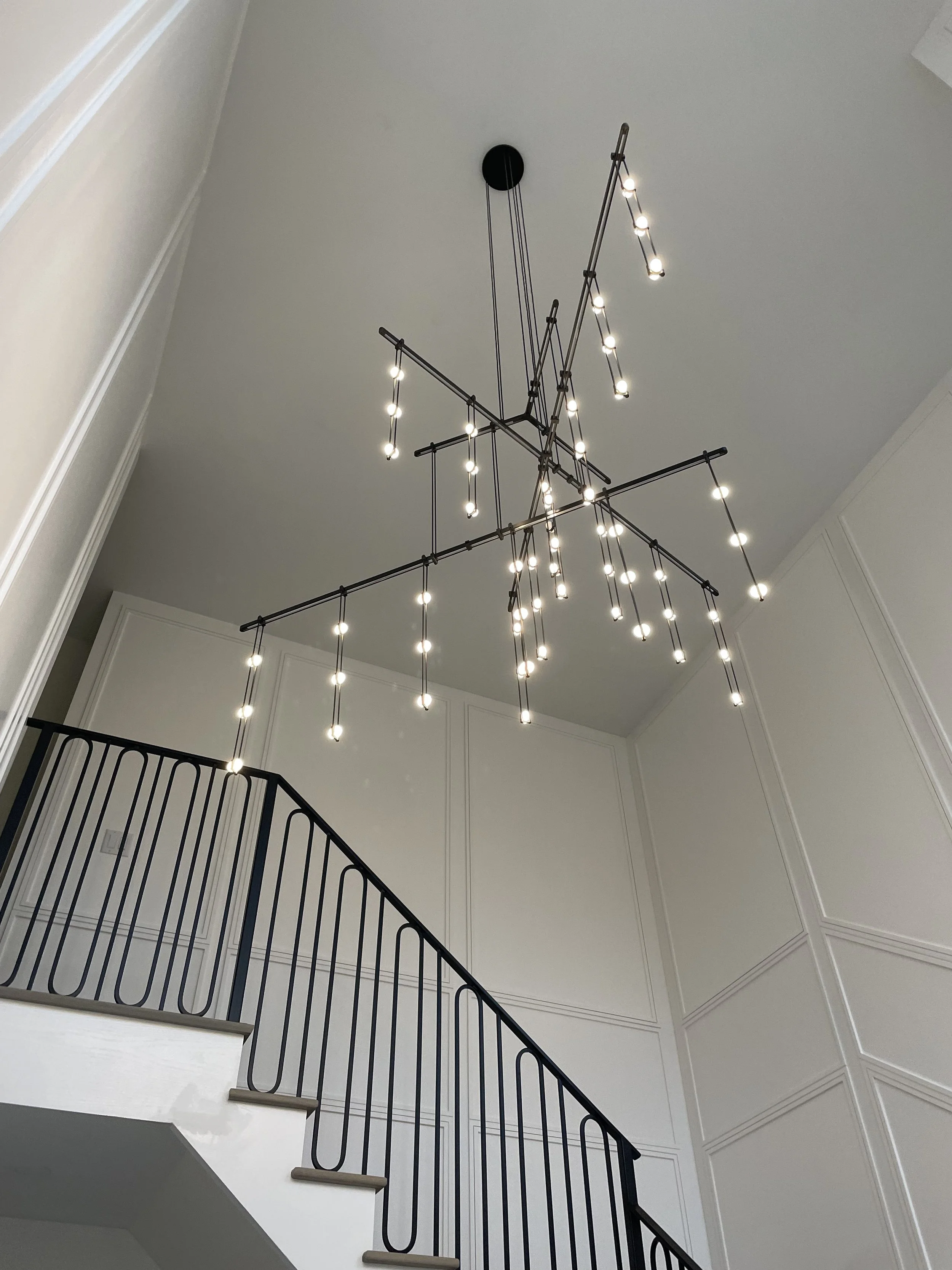 Modern chandelier with multiple small lights hanging from a black metal structure above a staircase with a black metal railing in a bright, white-walled interior.