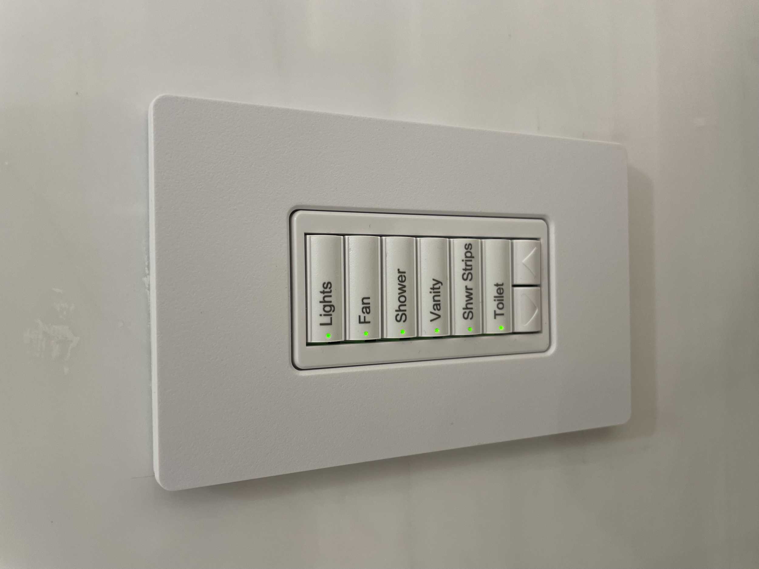 Indoor light switch panel with multiple switches labeled for different functions like lights, fan, vanity, shower strips, and toilet, all with green indicator lights.