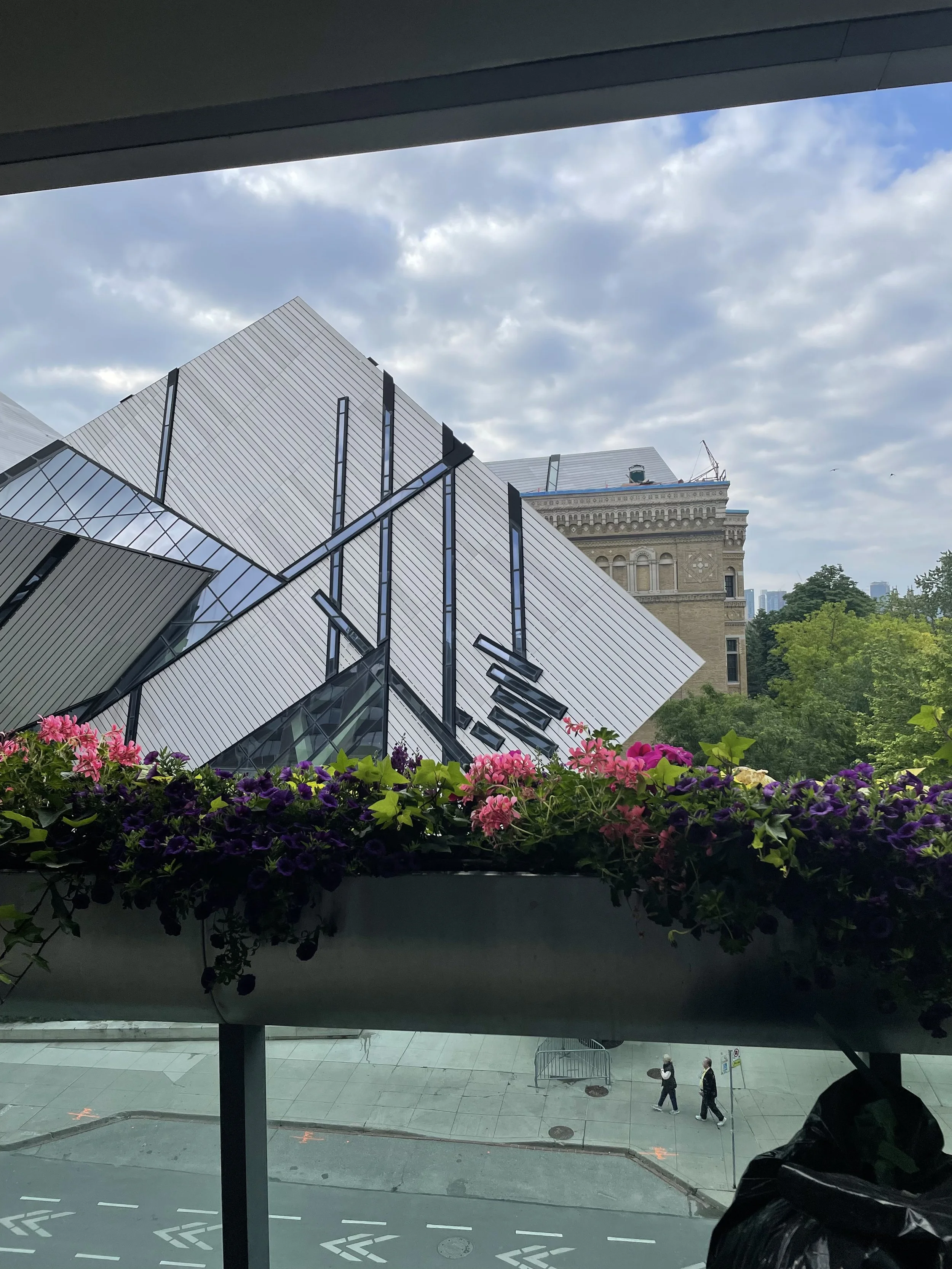 View from a window showing a modern building with angular metal and glass architecture, a historic building in the background, a flower box with pink, purple, and yellow flowers, and a street with pedestrian walkways and a few people walking.
