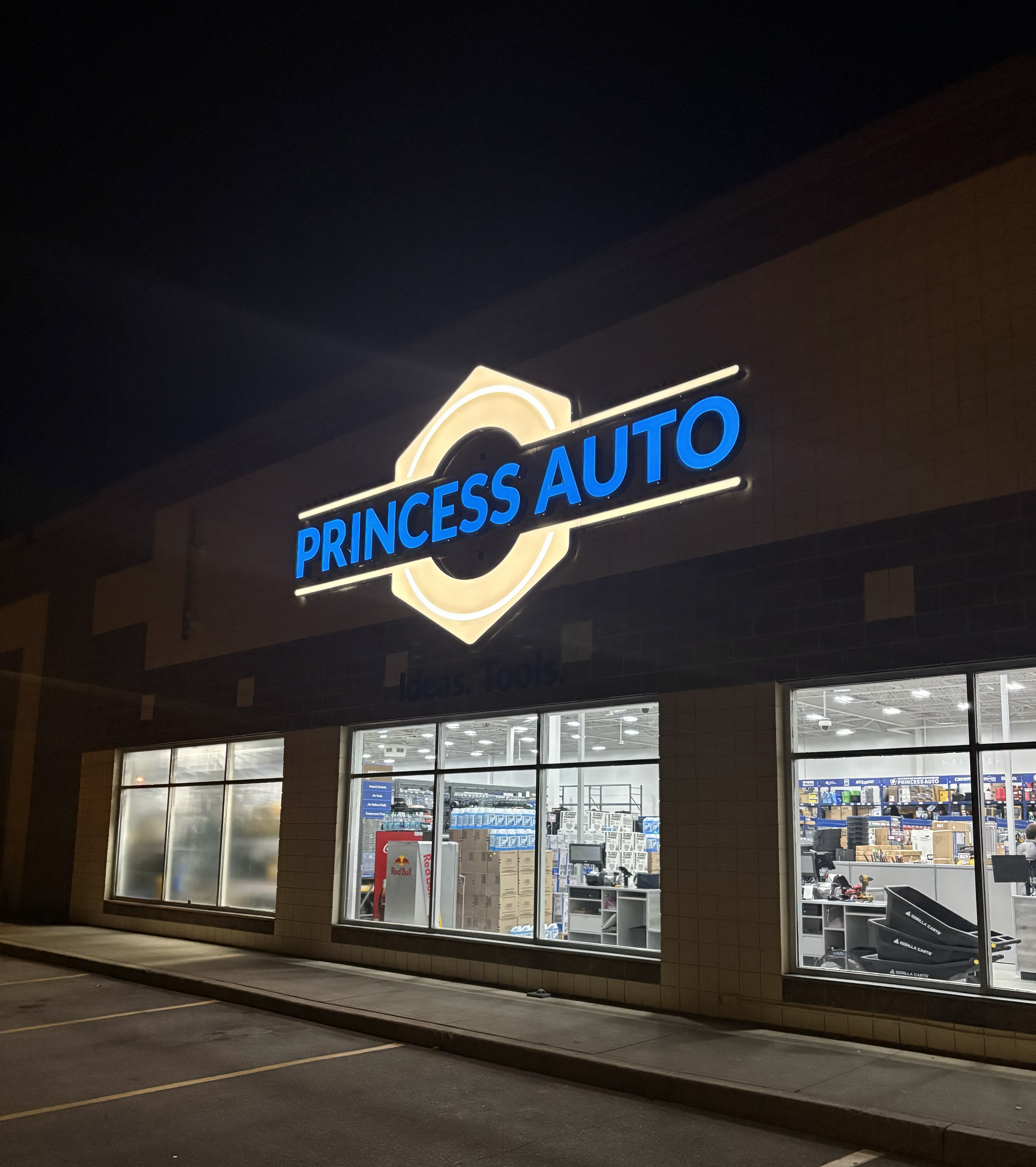 Nighttime view of a store front with illuminated sign that reads "PRINCESS AUTO" in blue letters and a logo above it. The shop interior is visible through large glass windows, displaying various automotive and hardware items, with a vending machine a