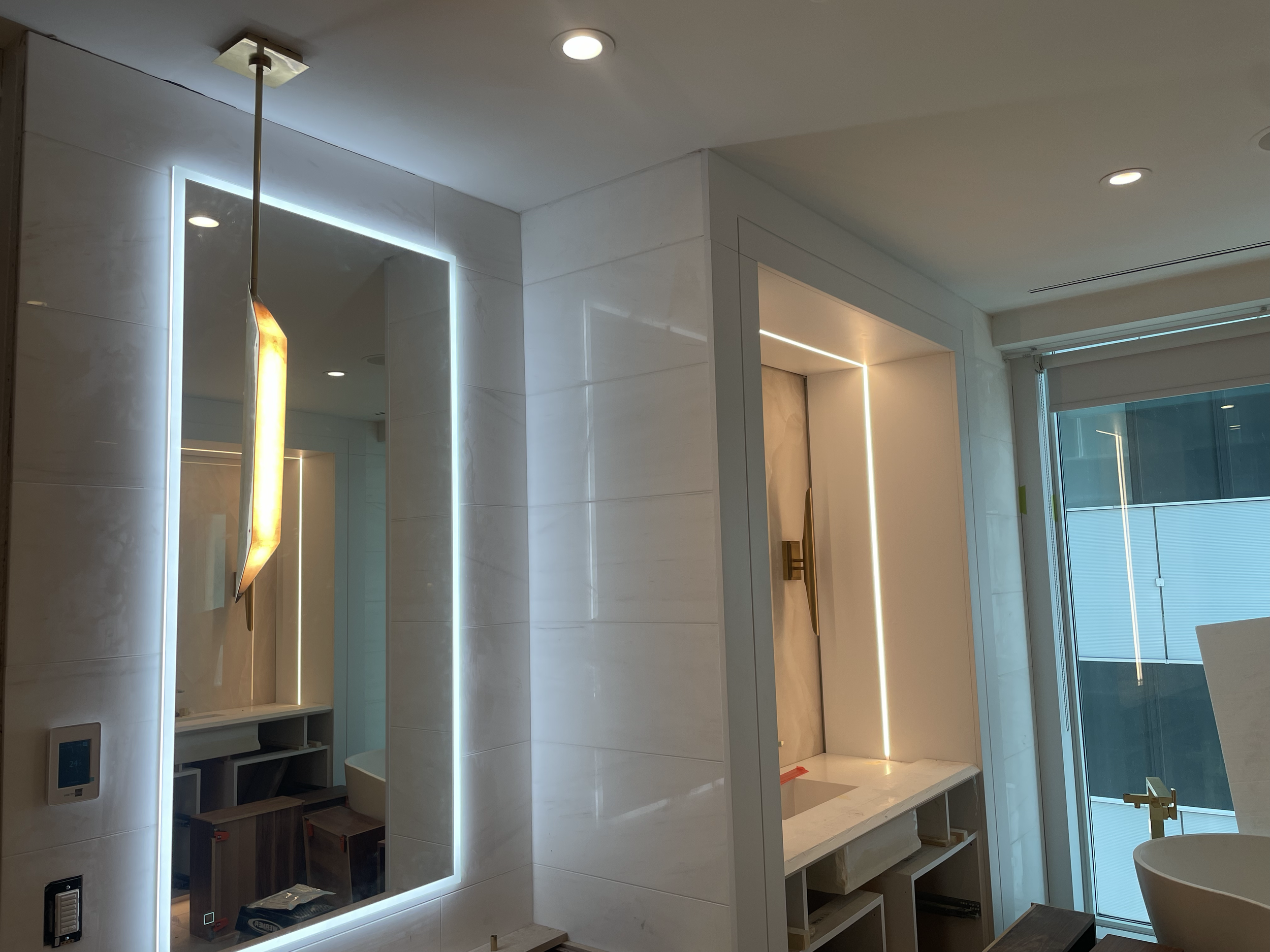 A modern interior with white tiled walls, vertical LED lighting strips, and a ceiling with recessed lights. There are some unfinished furniture pieces and construction materials on the floor.