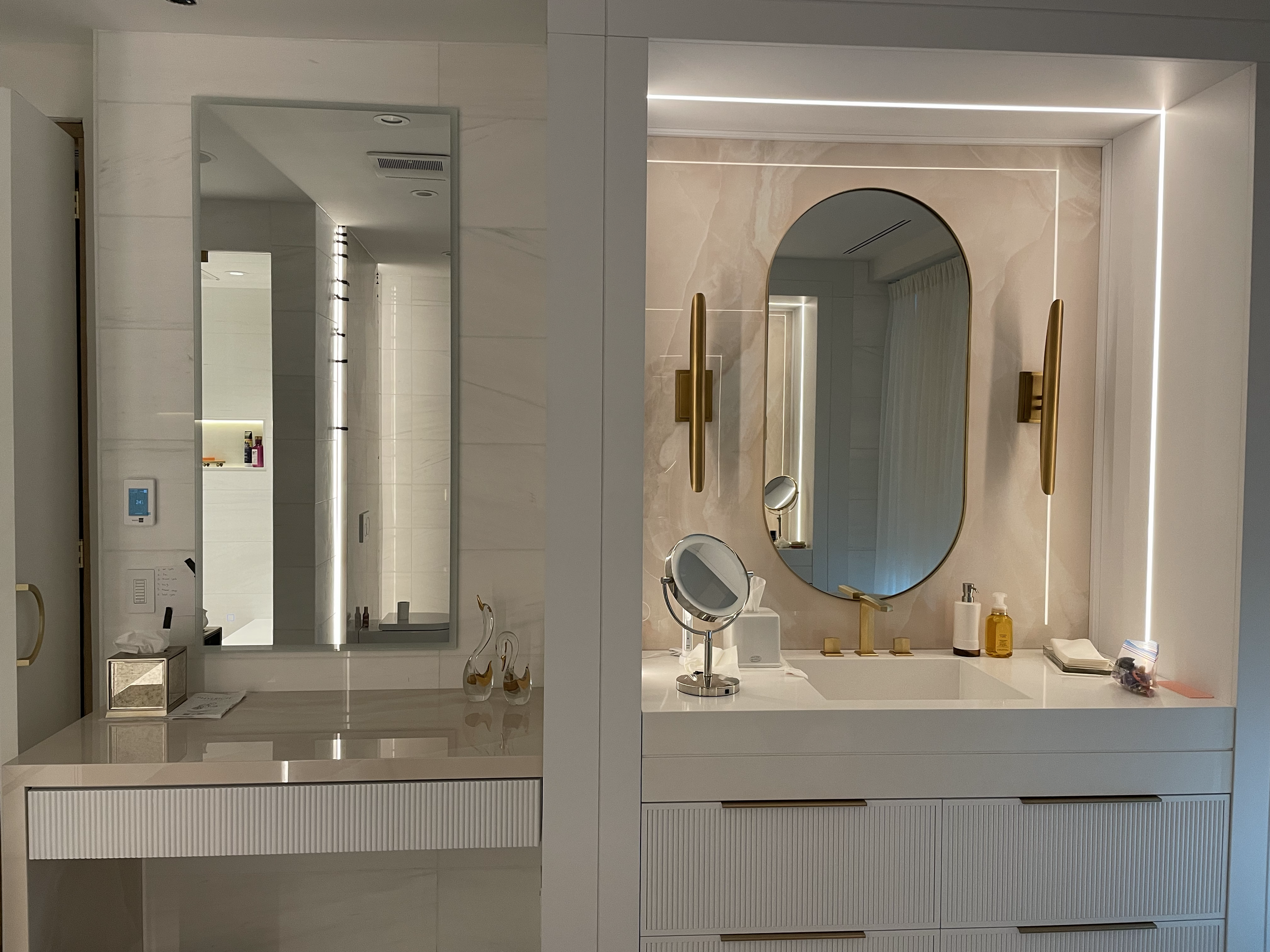 The image shows a modern bathroom vanity with a large round mirror, golden handles, a small circular magnifying mirror, various toiletries including soap, lotion, and a makeup remover, framed pictures, and a light-colored marble backsplash. Reflectio