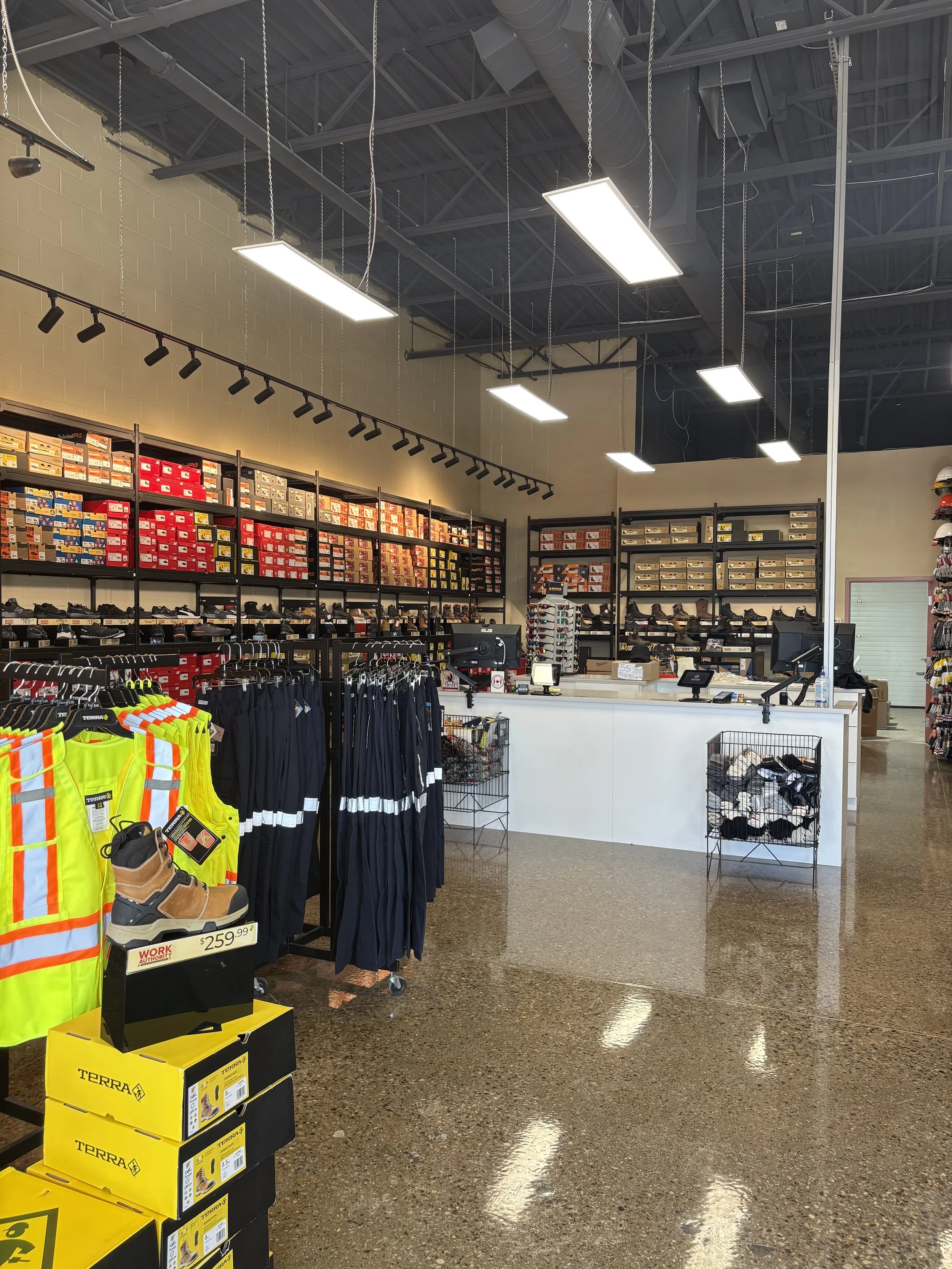 Interior of a retail store with shelves stocked with shoeboxes, clothing on racks, and a checkout counter. Fluorescent lights hang from a black ceiling with exposed ductwork. The floor is a glossy, speckled surface.