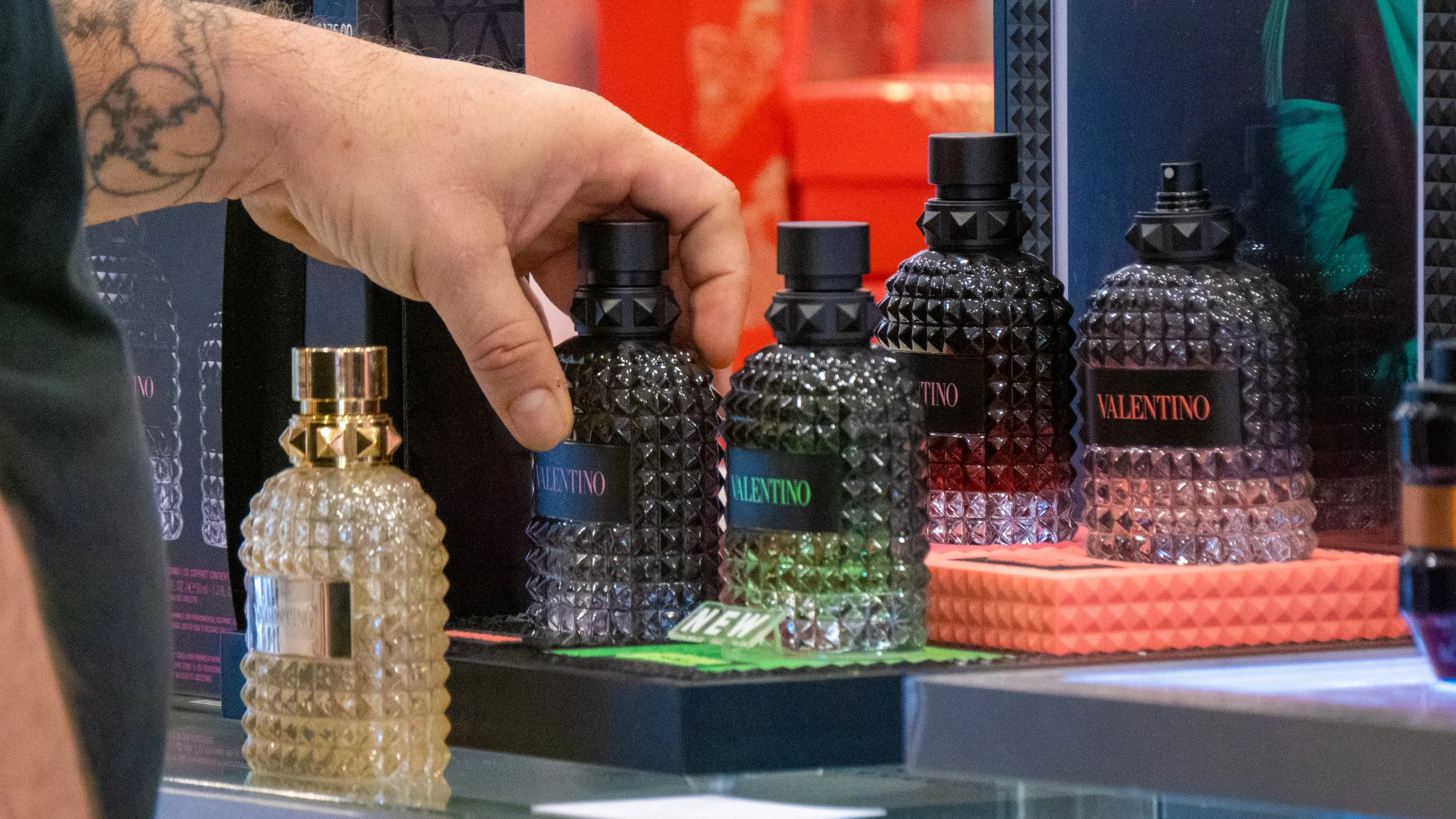 A person's hand with a tattoo selecting a black designer perfume bottle at a display of Valentino perfumes. Several similarly styled bottles and other fragrances are on a table.