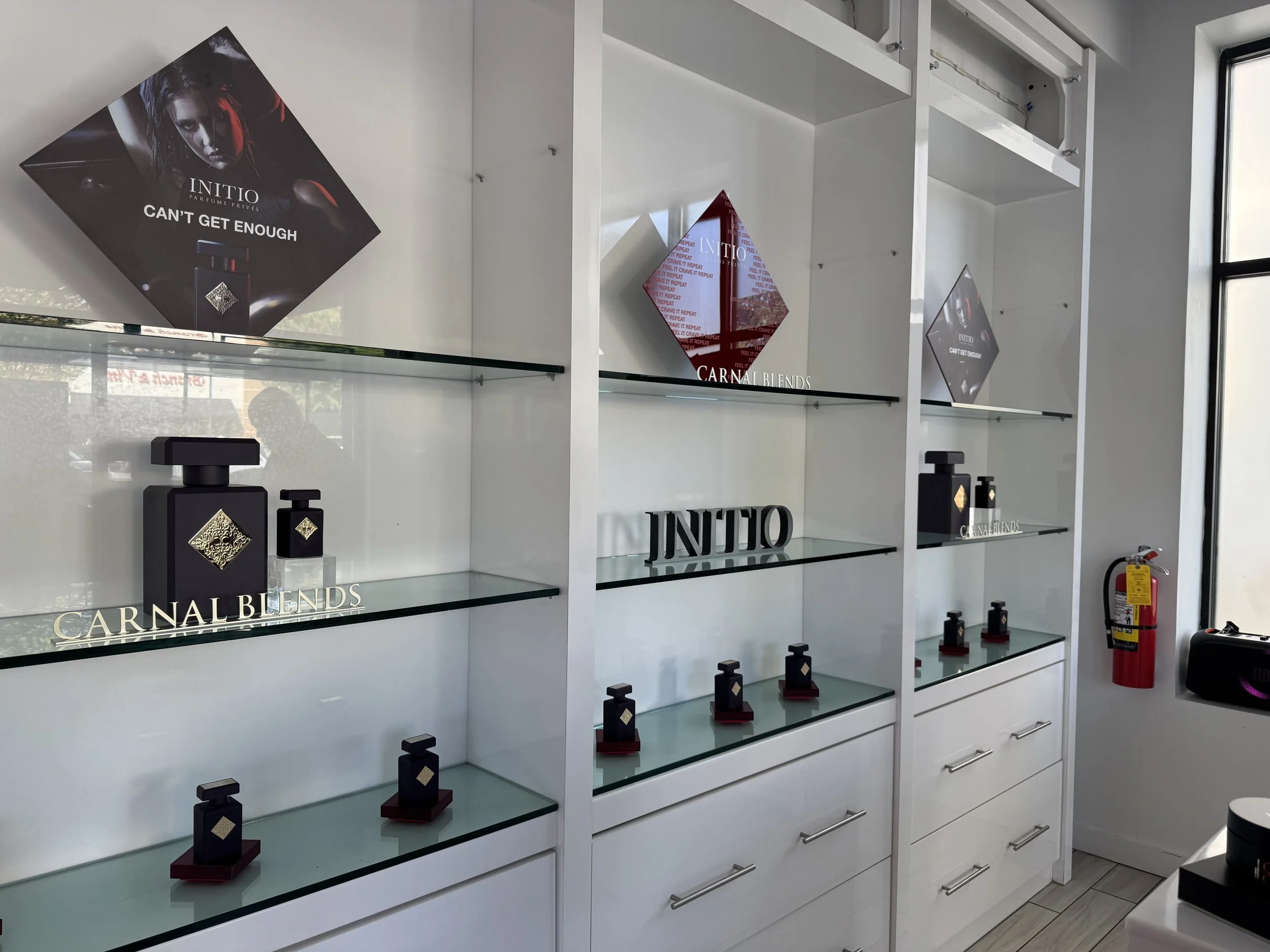 Display shelves featuring perfume bottles, promotional posters for Initio perfume, and a fire extinguisher in the corner of a retail store.