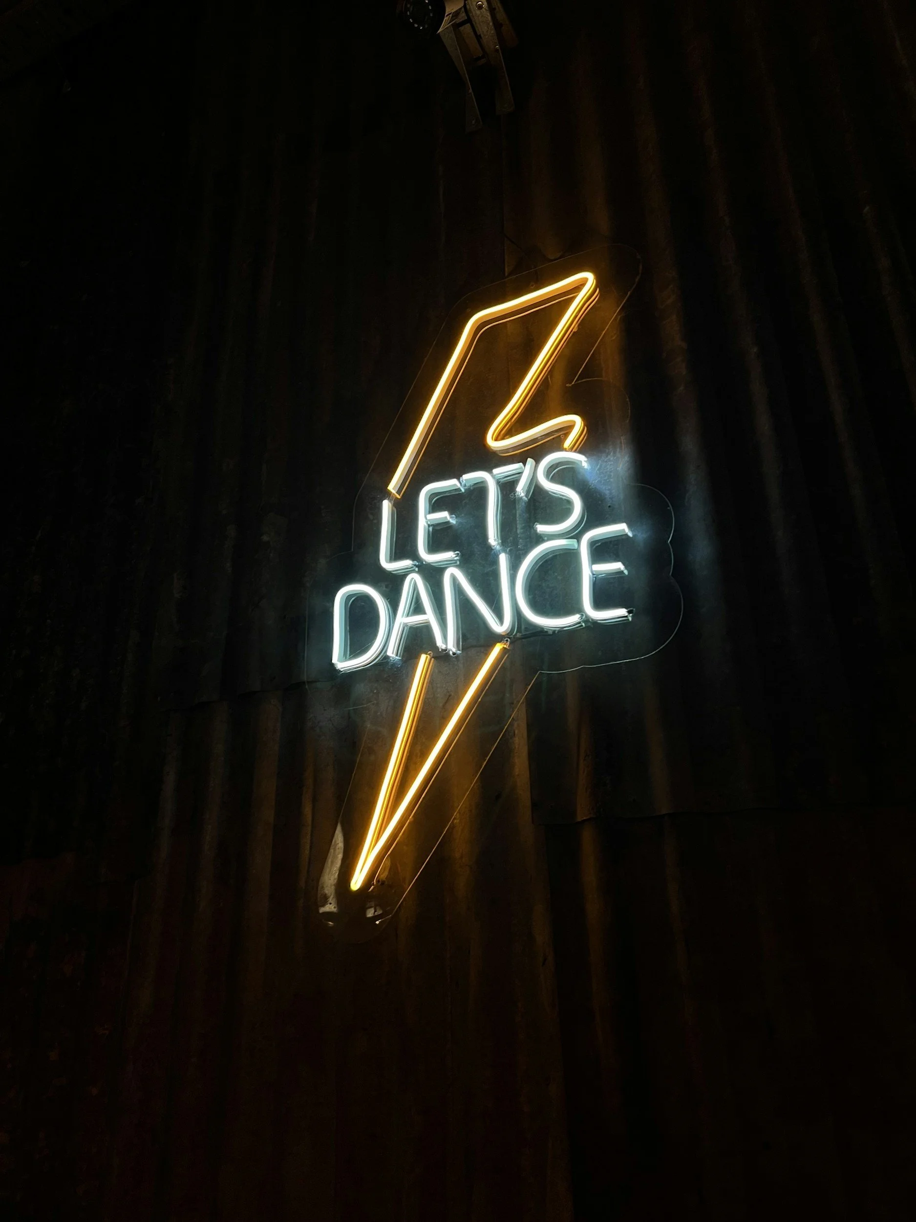 Neon sign reading 'Let's Dance' with a lightning bolt design, illuminated in yellow and white against a dark background.