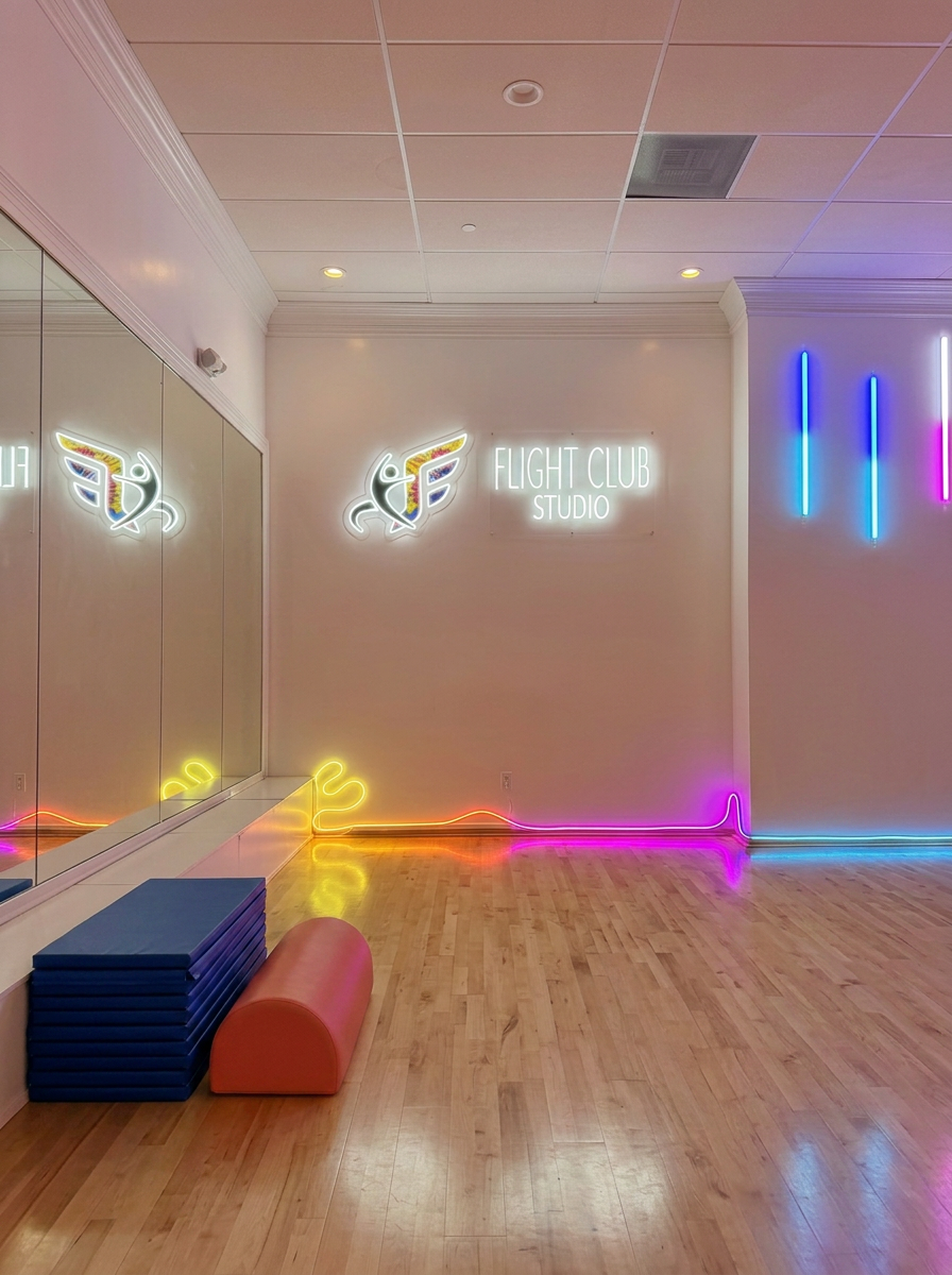 A brightly lit fitness studio with neon lights, including a sign that says 'Flight Club Studio' and decorative neon wings. There are mirrors on one wall, and a blue exercise mat with an orange half-cylinder foam roller on the wooden floor.