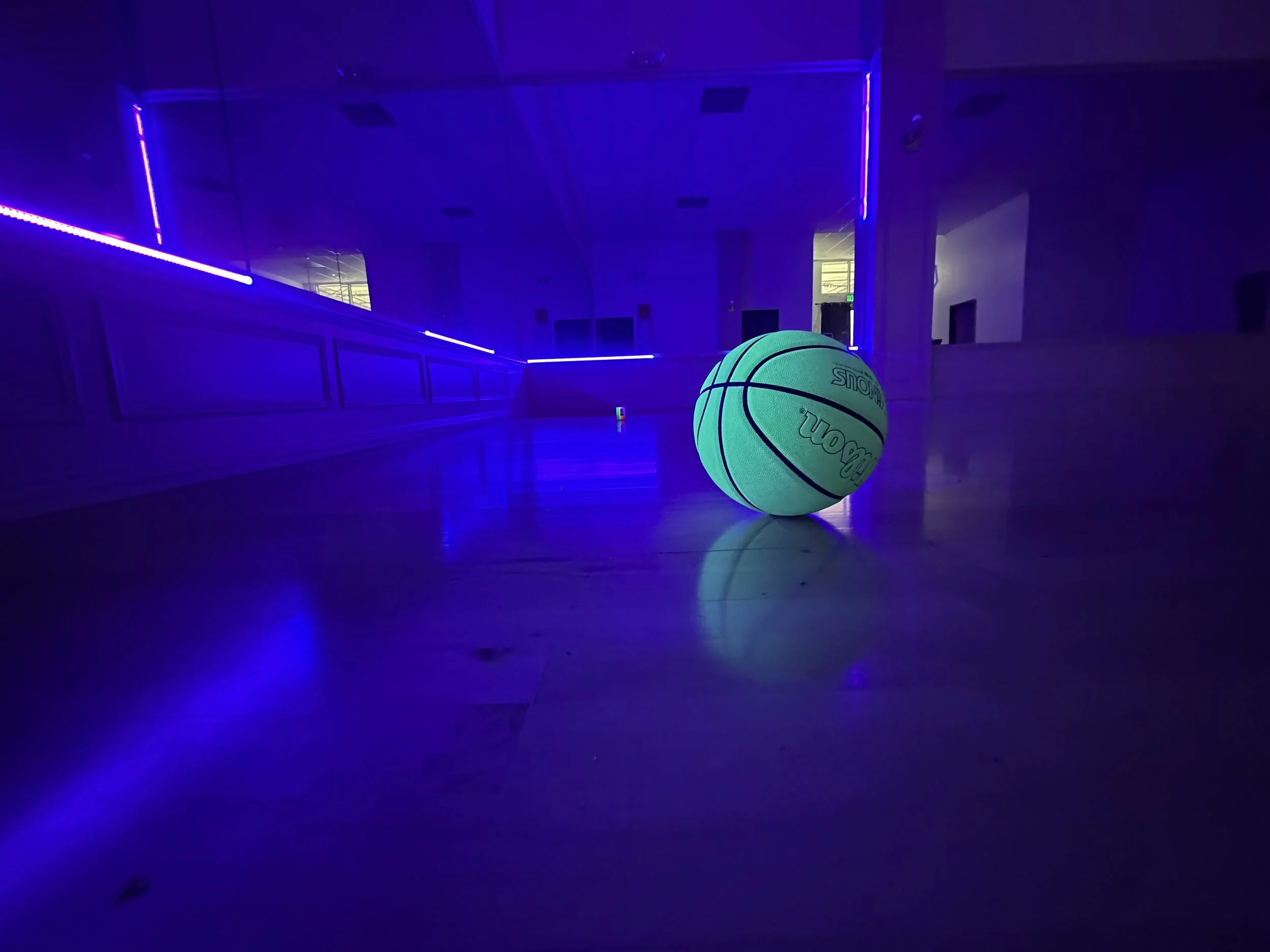 A glowing green basketball on a polished wooden floor in a dark room illuminated by blue and purple neon lights.