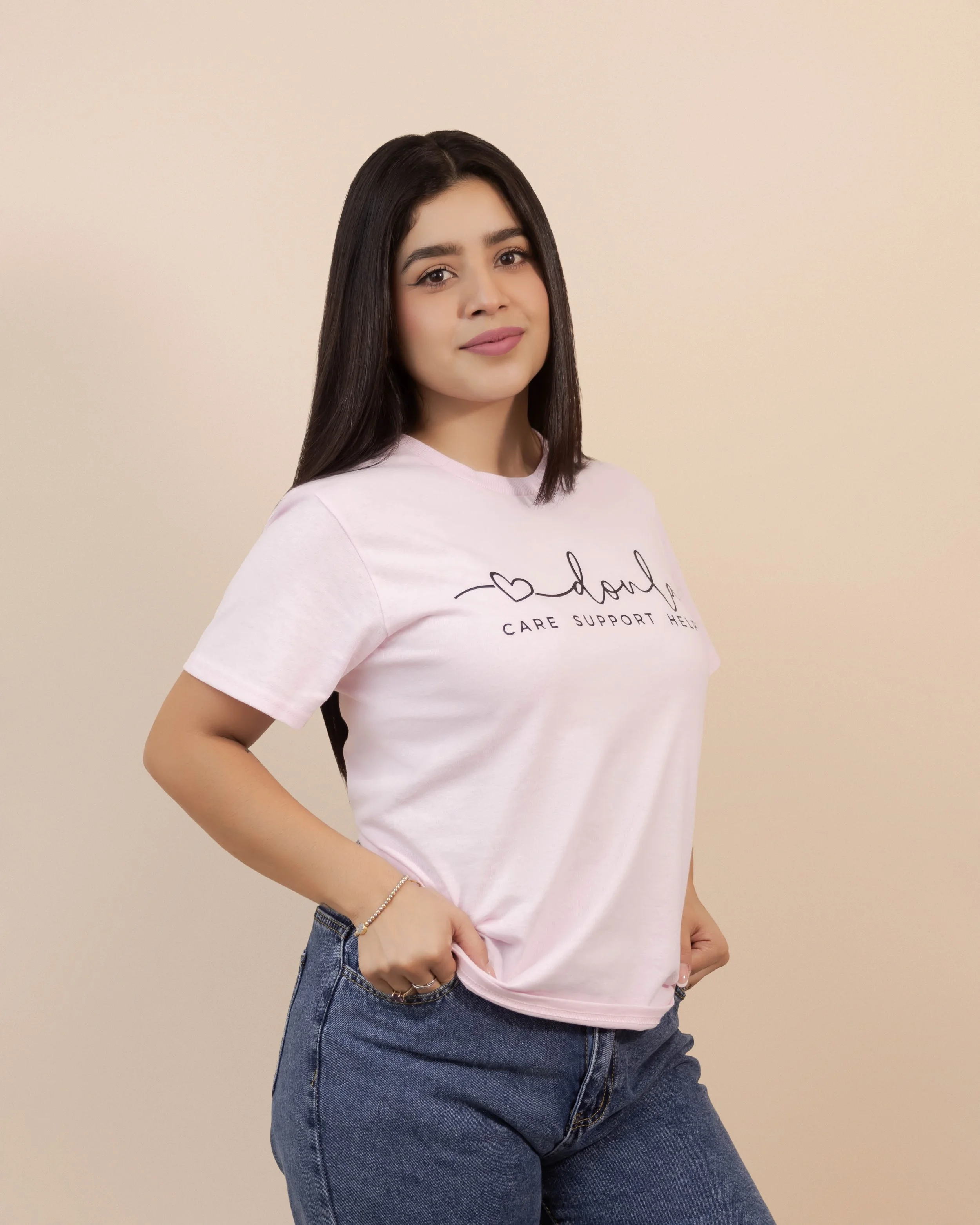 A young woman with shoulder-length dark hair, wearing a light pink T-shirt with black text and dark jeans, standing against a plain beige wall, posing with her right hand on her hip and smiling softly.
